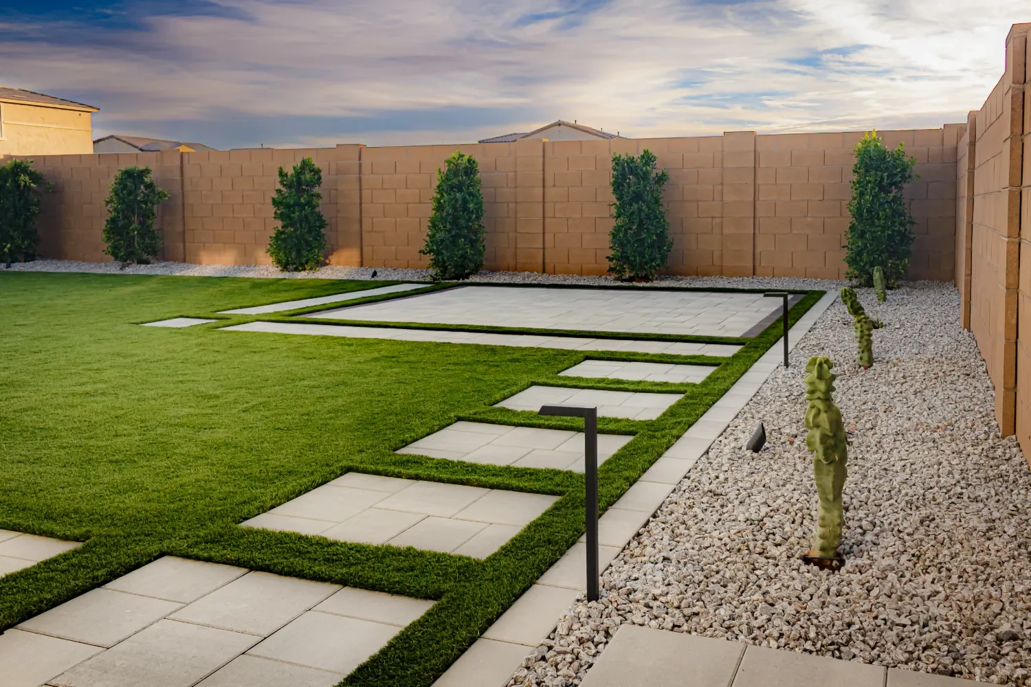 Modern backyard with a walkway made of large square tiles and grass in between, a well-maintained lawn, tall green trees along a beige brick wall, and desert plants on the gravel side.