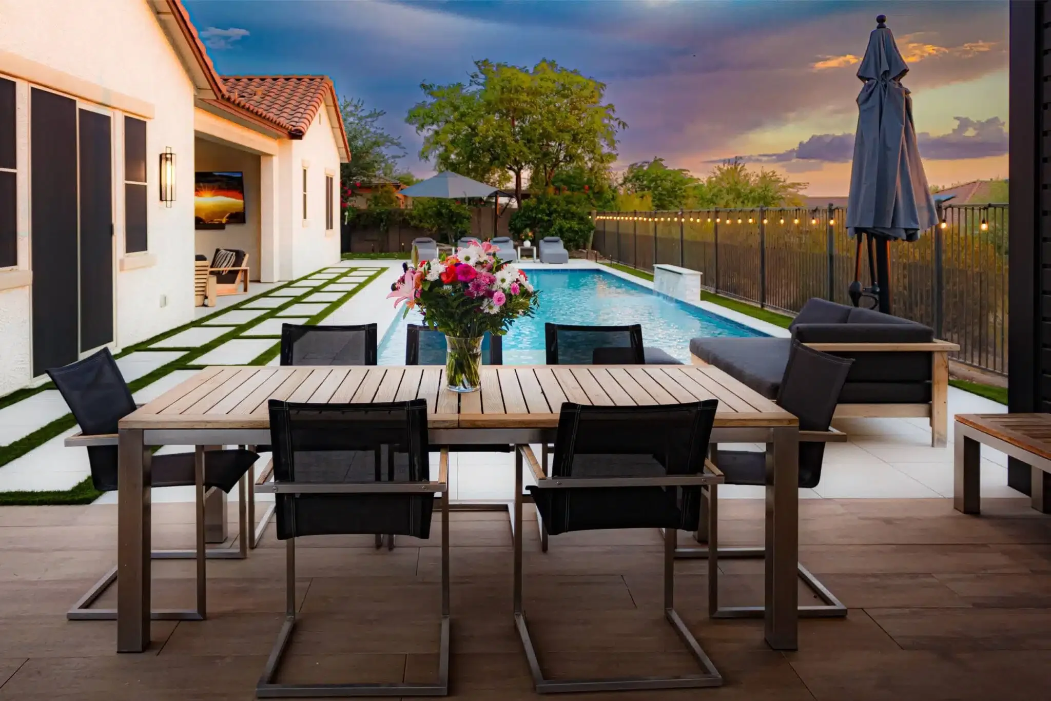 A backyard at sunset featuring a swimming pool, outdoor dining table with six chairs and a flower arrangement, lounge chairs near the pool, with a tree and fence in the background in Queen Creek, AZ