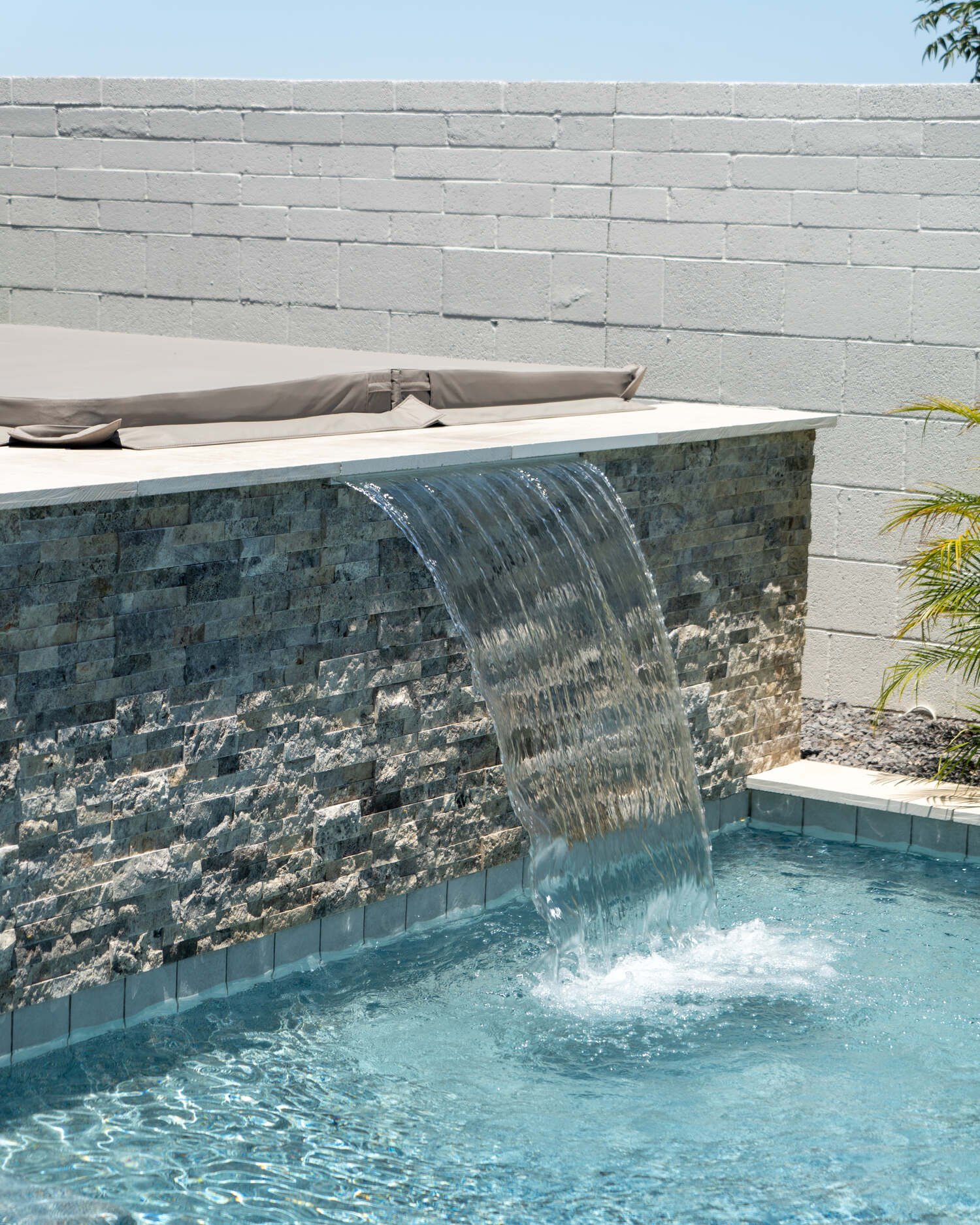 Water feature with waterfall over a stone wall into a swimming pool, with a white ledge and umbrellas on top in Arcadia, AZ