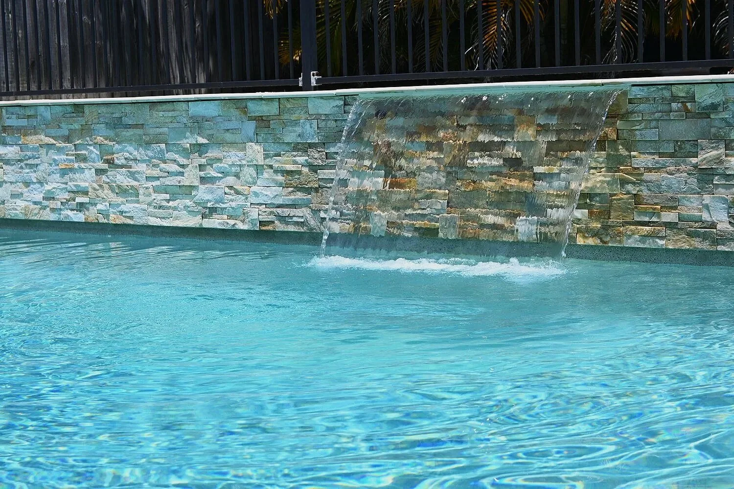Swimming pool with a waterfall feature cascading into the water, surrounded by a stone wall and a black metal fence in Paradise Valley, AZ