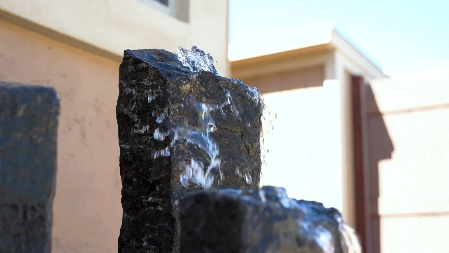 Close-up of black rocks with water flowing over them, outdoors with a building and fence in the background in Queen Creek, AZ