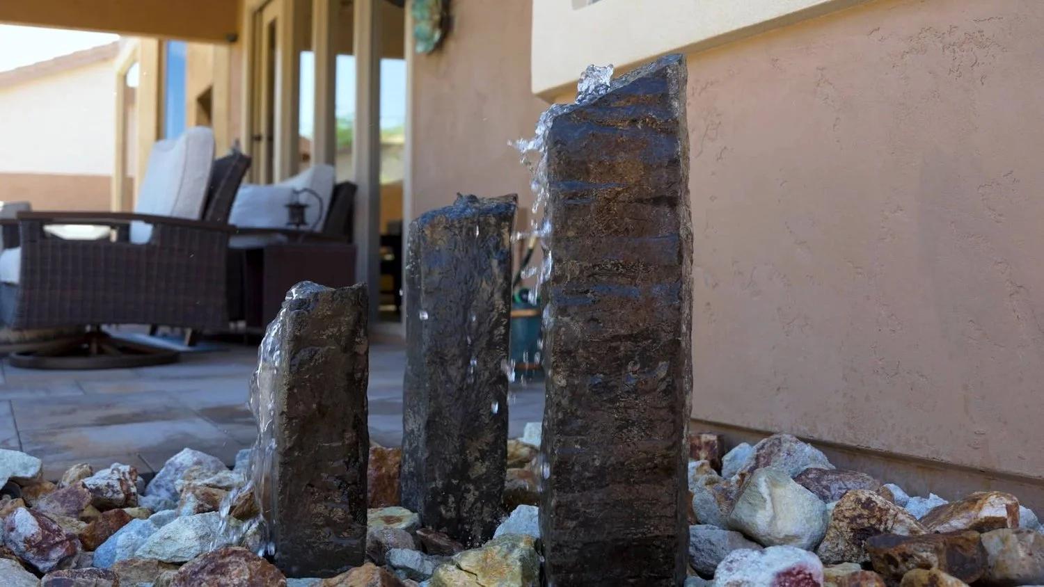 Close-up of a small outdoor water fountain with three dark stone pillars and water flowing down, surrounded by multicolored rocks on a patio with outdoor furniture in the background in Vistancia, AZ