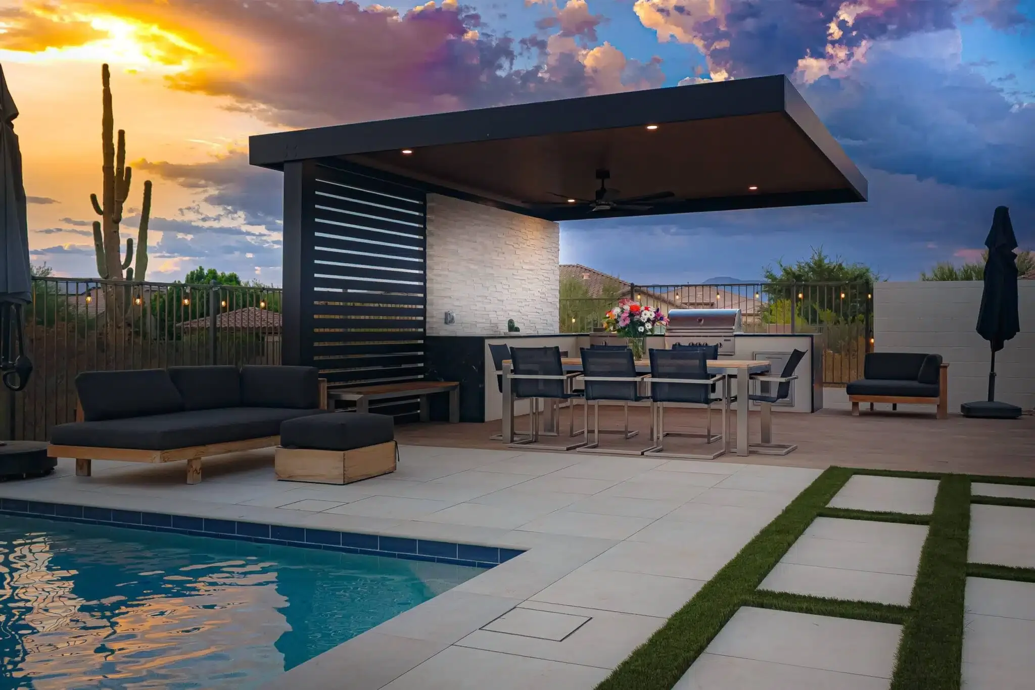 Modern outdoor patio area with a pool, black and wooden furniture, a dining table with chairs, a barbecue, a flower arrangement, and sun umbrellas, set against a sunset sky with clouds in Surprise, AZ