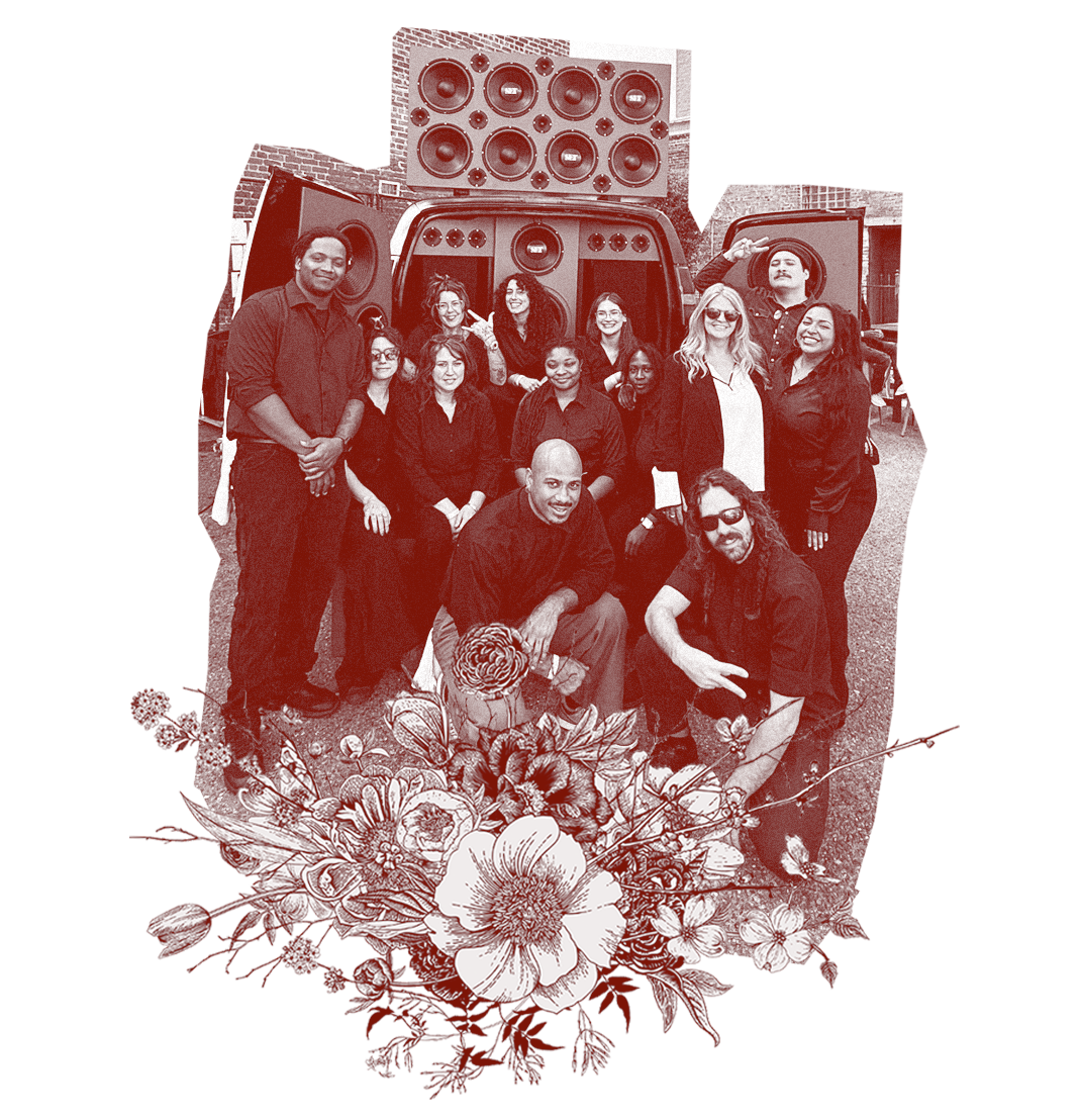 Group of people posing in front of a large speaker system, with some standing and some kneeling, outdoors.