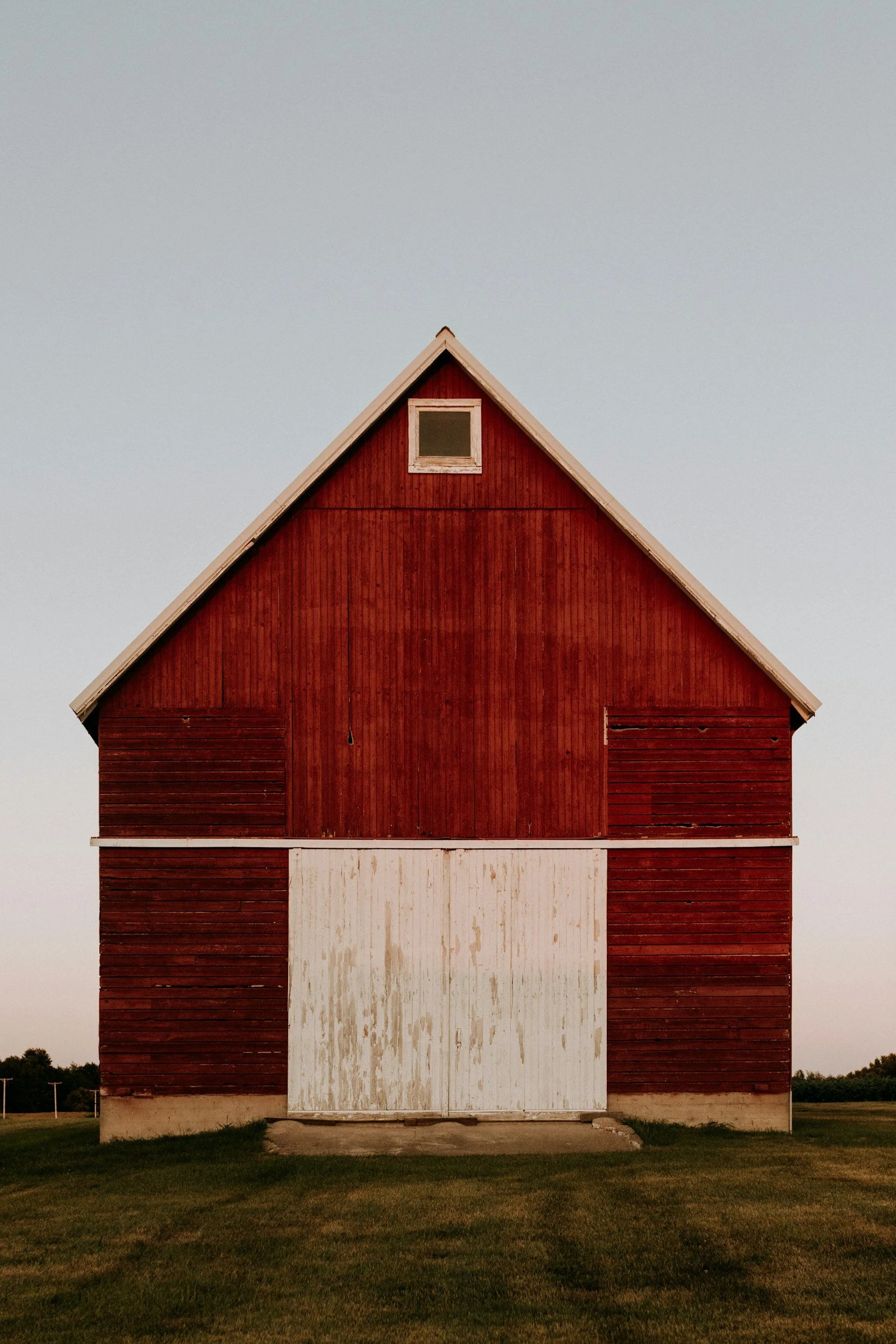 A large red wooden barn with white trim and a white sliding door, situated on a grassy field with a clear sky in the background.