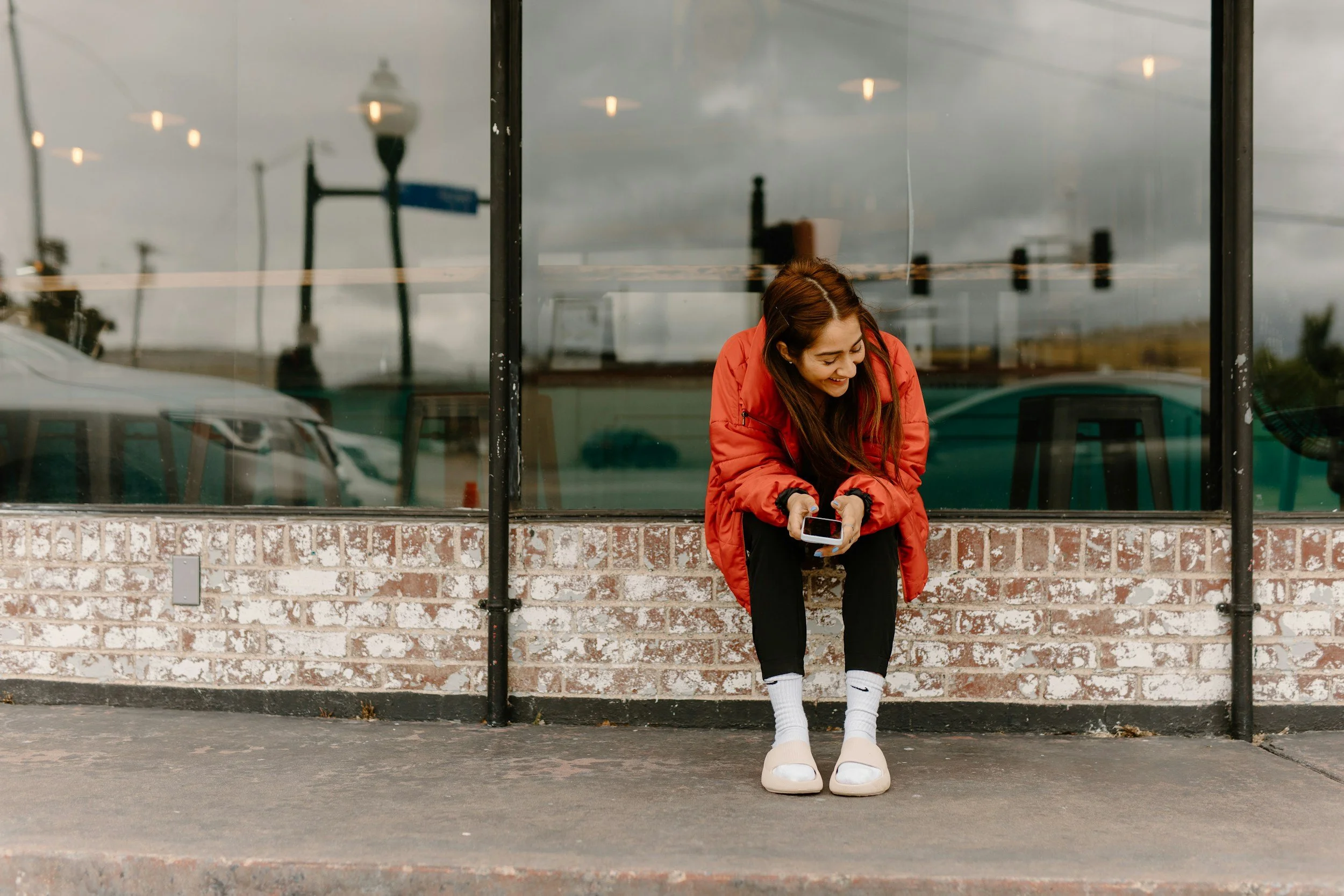 A young woman with long brown hair, wearing a red jacket, black pants, white socks, and sandals, sitting on a curb outside a glass-front building, looking down at her phone and smiling.