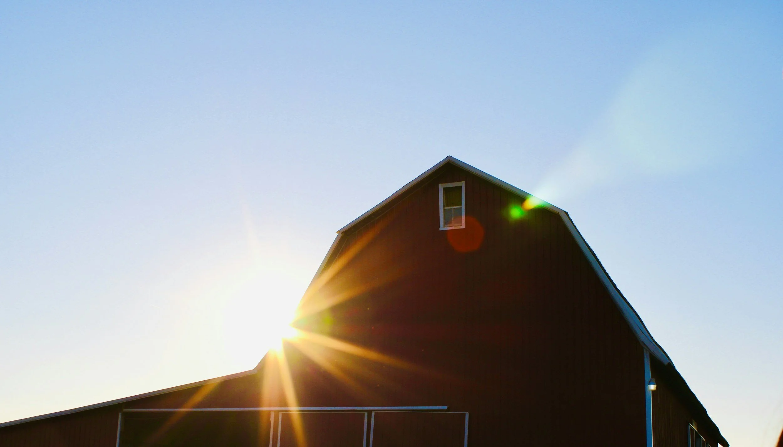 A red barn silhouette with the sun setting behind it, creating a bright flare and sun rays against a clear blue sky.