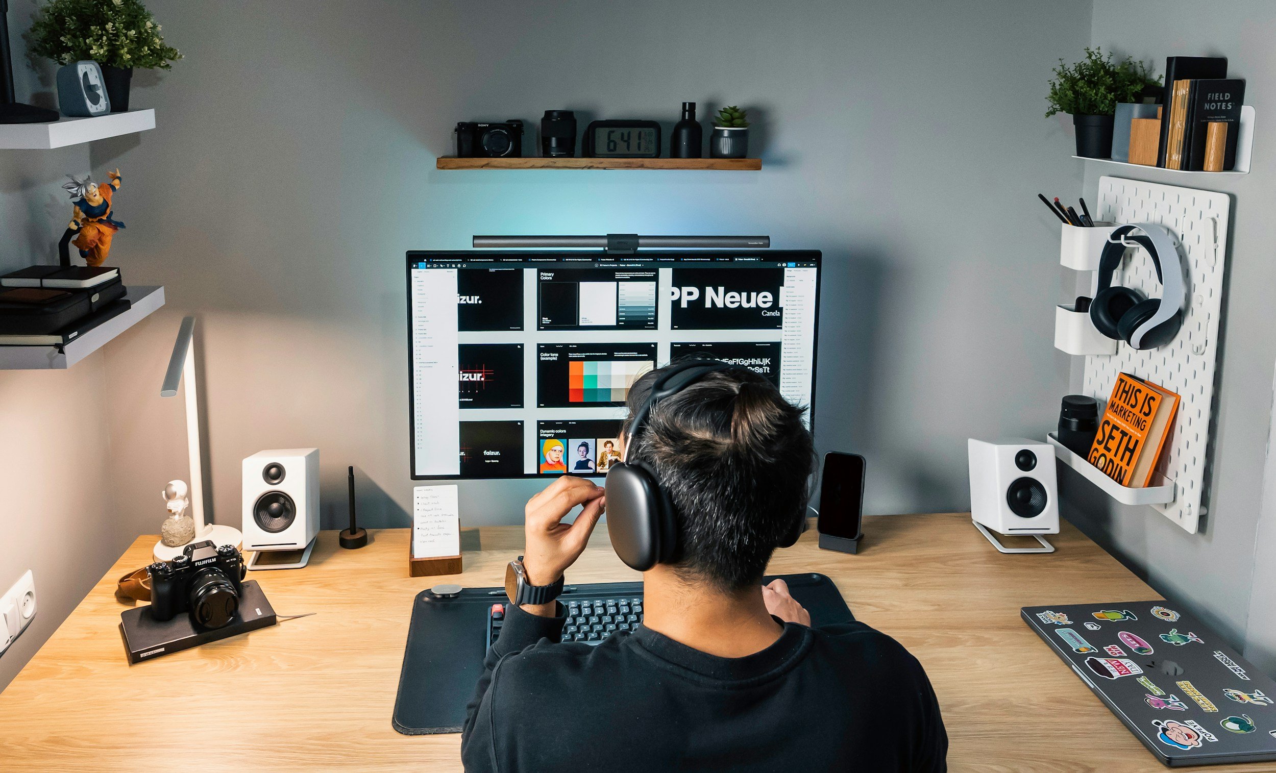 A person sitting at a desk with headphones on, looking at a computer screen displaying a digital design software. The desk has a camera, speakers, a smartphone, a keyboard, and a laptop covered with colorful stickers. The background includes shelves with books, plants, and camera gear, and a pegboard with headphones and books.
