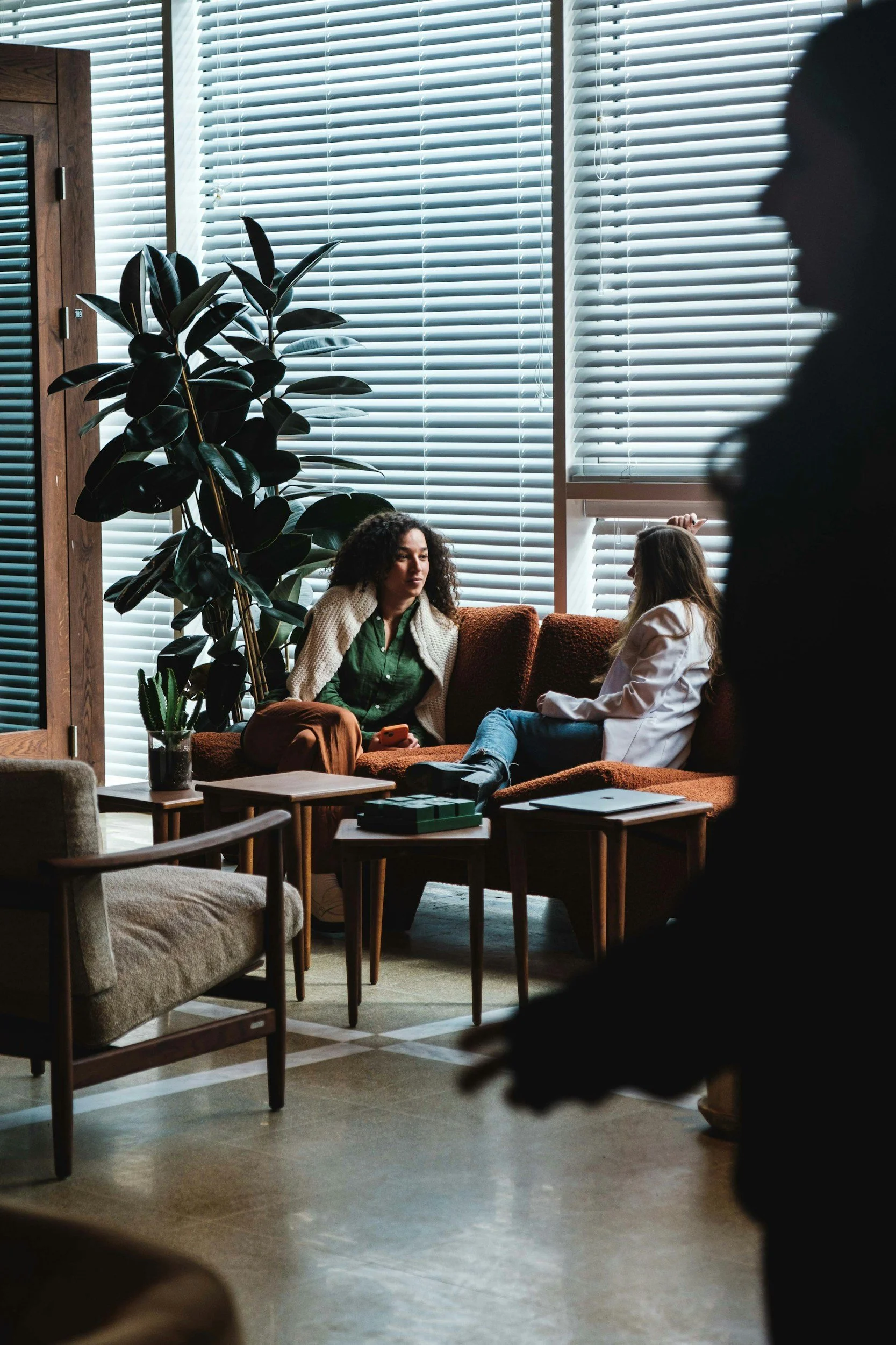 Two women sitting on a brown couch having a conversation, with one woman holding a smartphone, in a room with large windows, blinds, indoor plants, and wooden furniture.