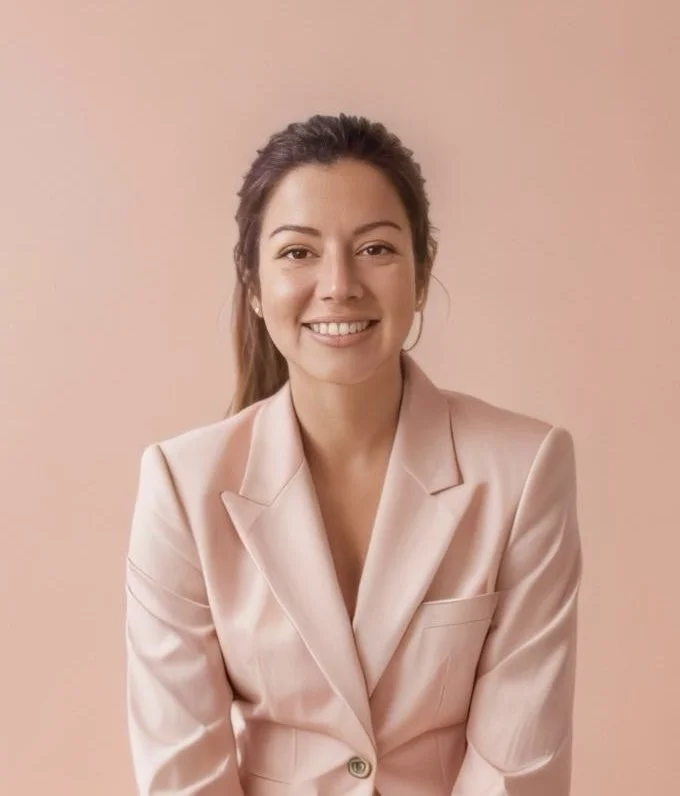 A smiling woman with brown hair tied back, wearing a light pink blazer, standing against a matching pink background.