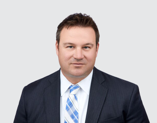 Professional man in a dark suit with a light blue and white striped tie, facing forward against a plain background.