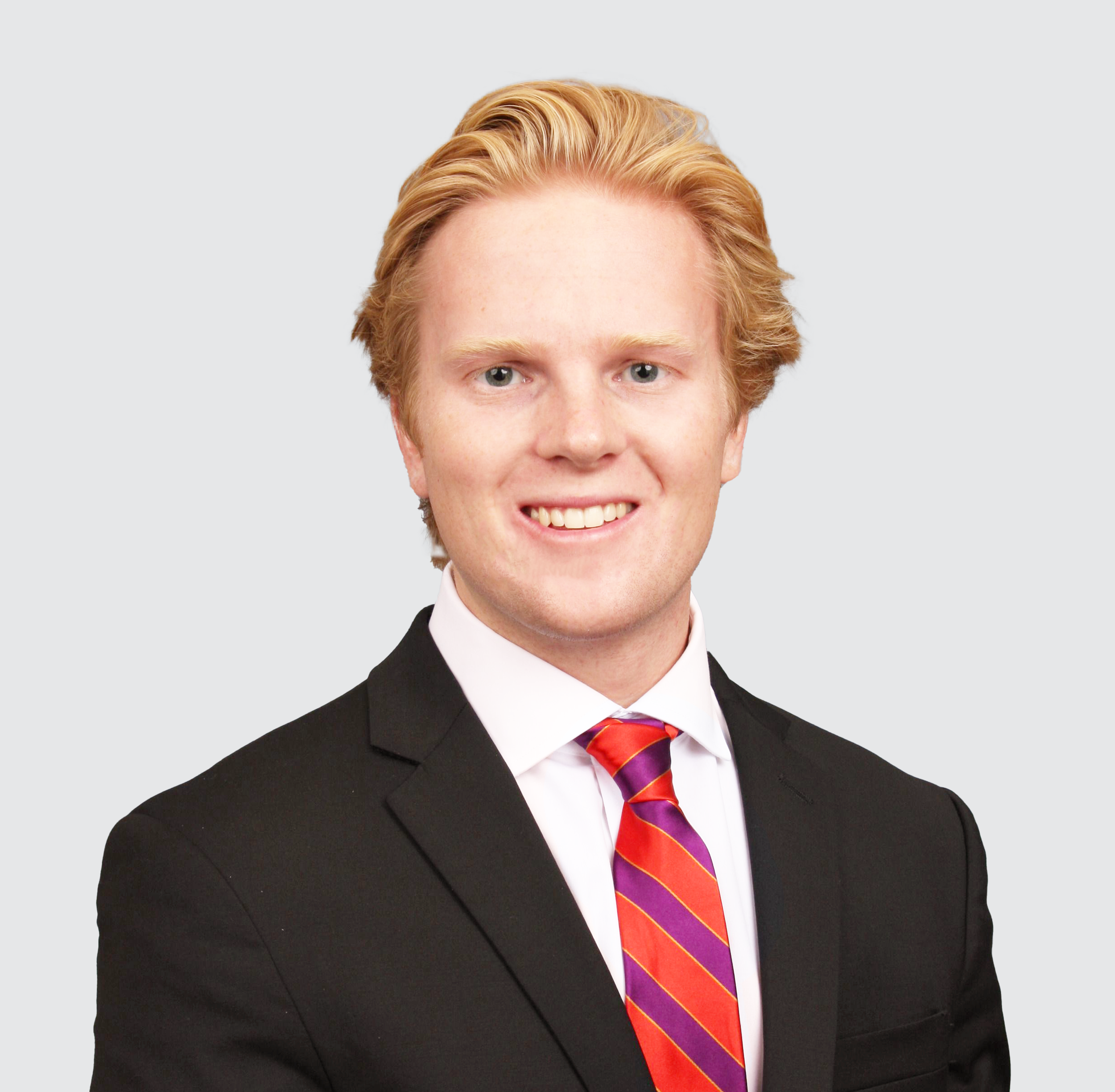 Portrait of a young man with blond hair, blue eyes, wearing a black suit, white shirt, and a striped red and purple tie, smiling against a plain light gray background.