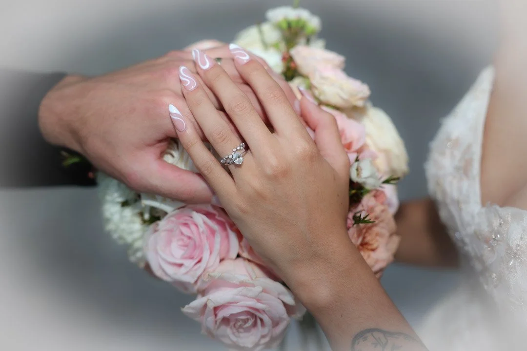 A bride and groom's hands resting on a bouquet of pink and white roses, showing a wedding ring on the bride's finger.