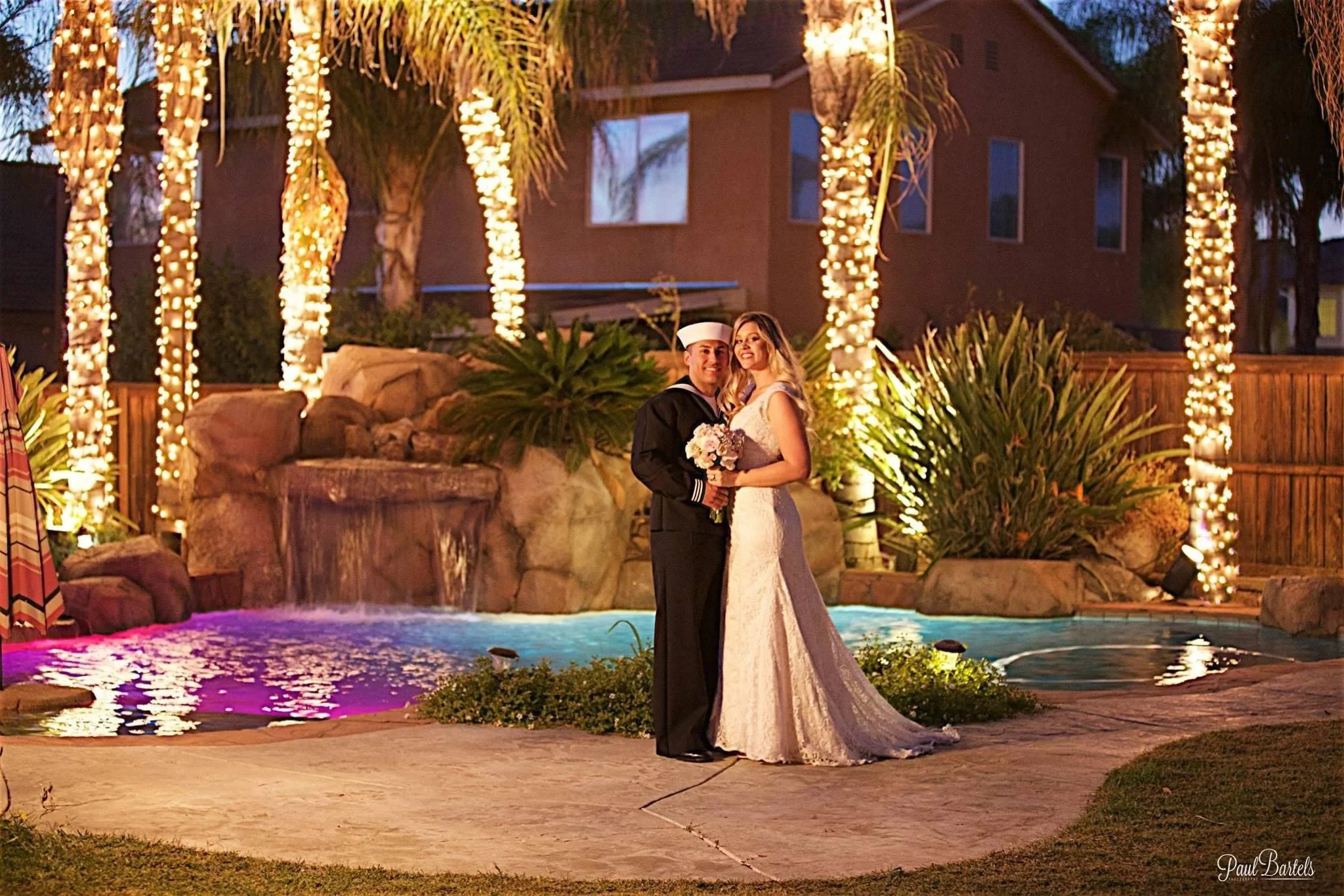 A newlywed couple stands in front of a backyard pool with waterfalls, surrounded by palm trees wrapped in string lights, during evening time. The groom is in a navy Navy sailor uniform, holding a bouquet, and the bride is in a white lace wedding gown, holding the bouquet with the groom. There are colorful underwater pool lights, decorative plants, and a wooden fence in the background.