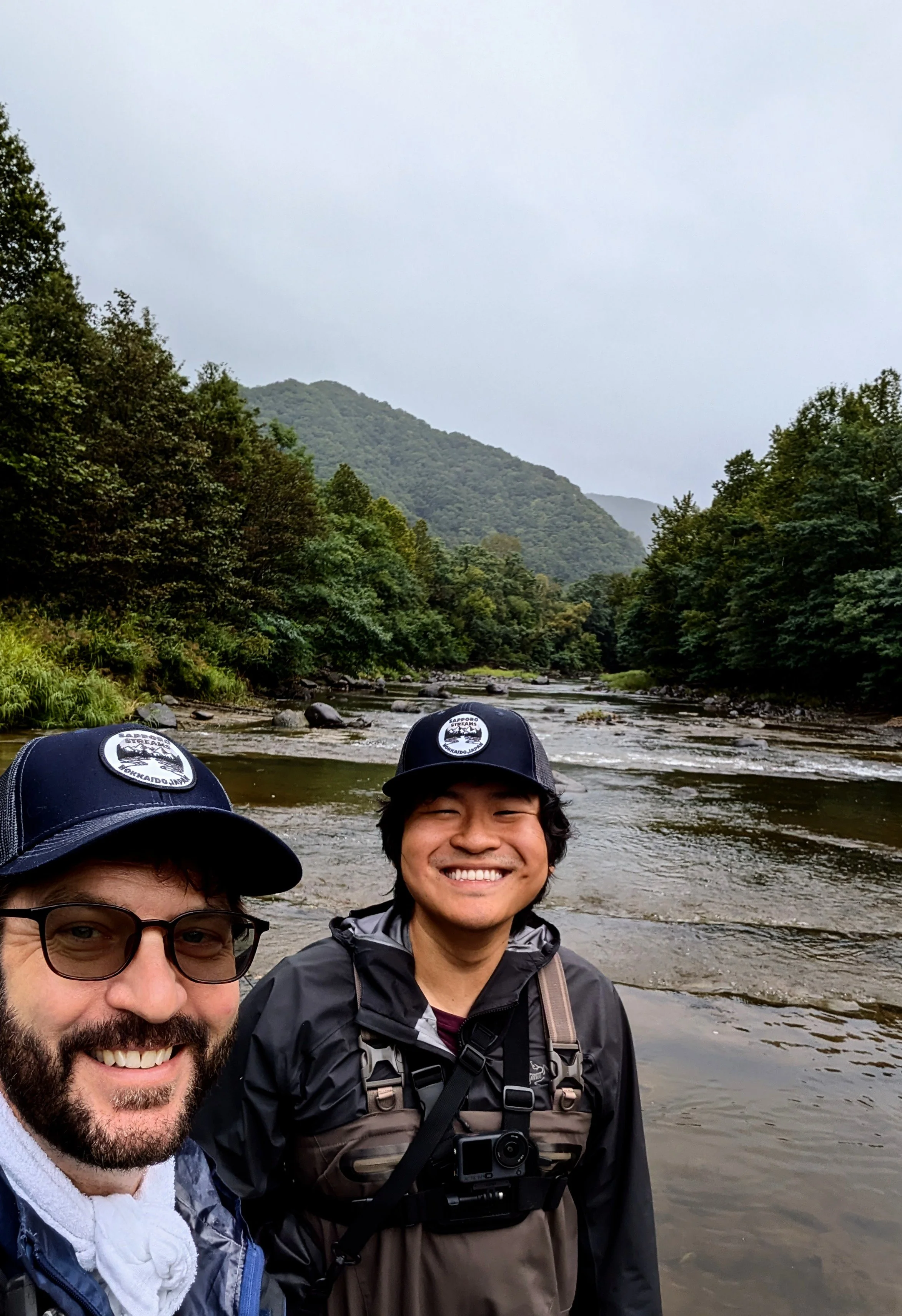 Two smiling men in a river surrounded by lush green trees and mountains under an overcast sky in Sapporo, Hokkaido Japan