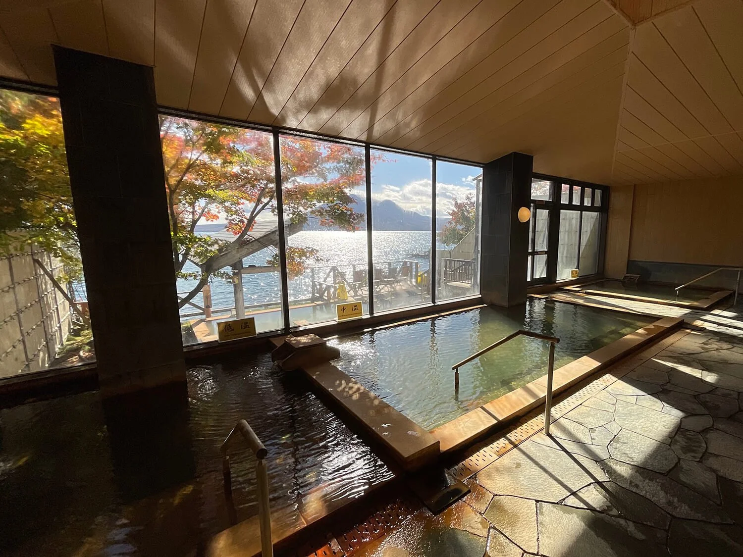 Indoor hot spring bath with large glass windows overlooking a lake and autumn trees outside, sunlight streaming in in Sapporo, Hokkaido Japan