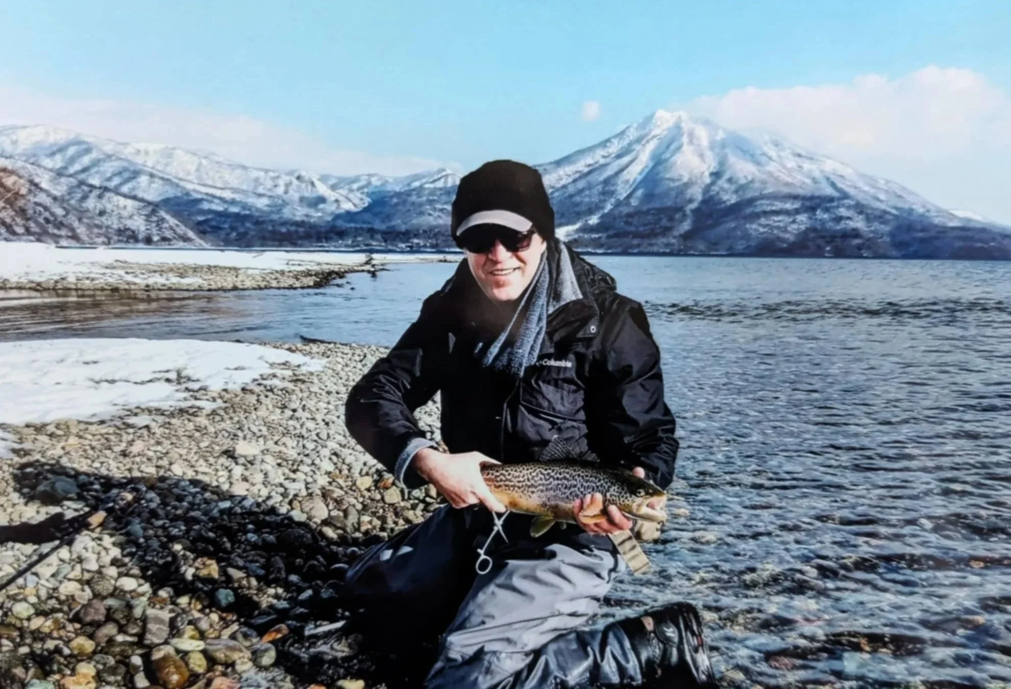 A person holding a large fish near a river with snow-covered mountains in the background in Sapporo, Hokkaido Japan
