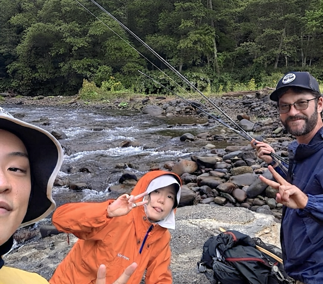 Three people near a river, one person holding fishing rods, two making peace signs, surrounded by rocks and trees in Sapporo, Hokkaido Japan