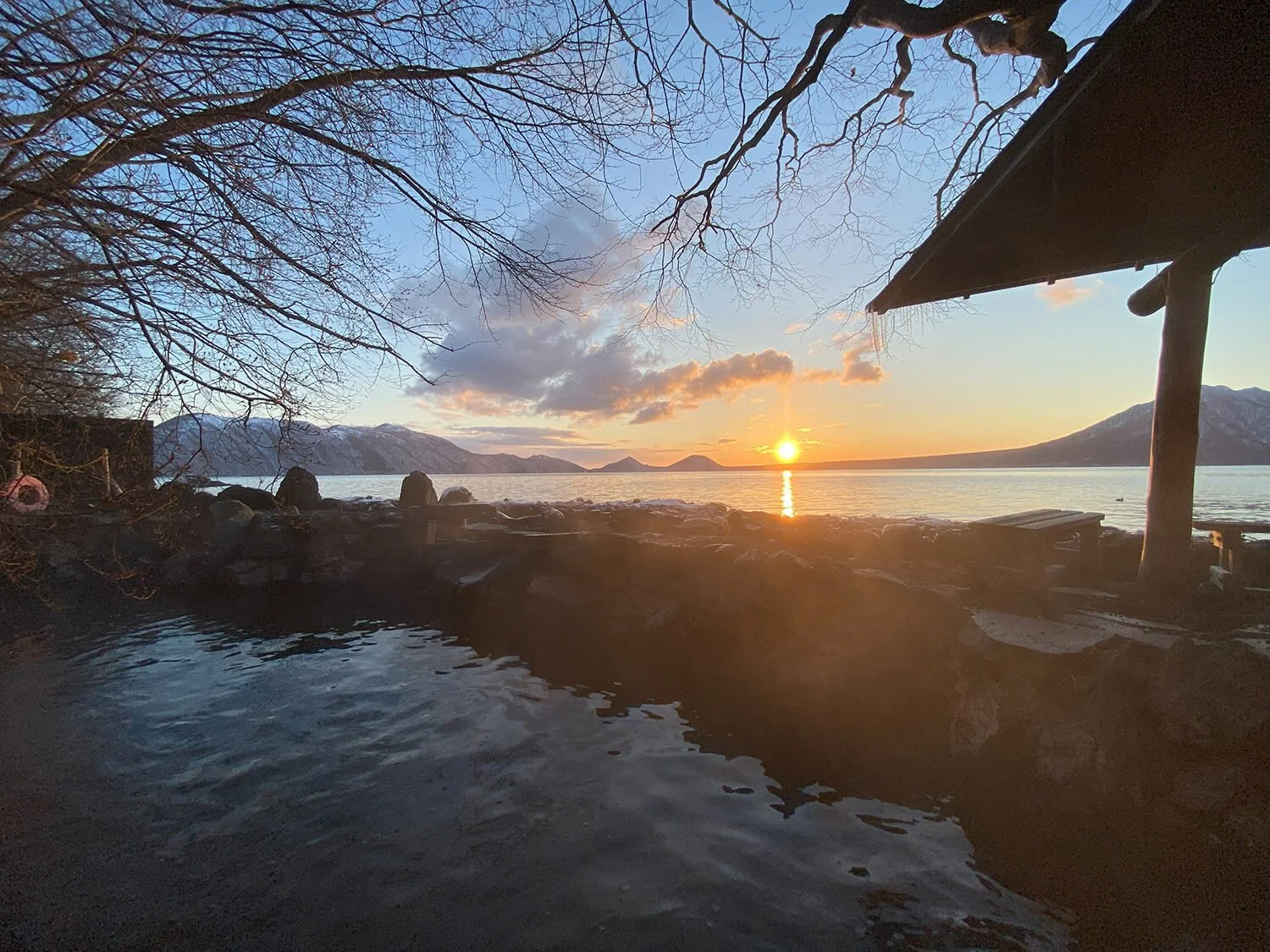 A peaceful sunset over a lake, with mountains in the background, bare trees in the foreground, and a small covered structure to the right.