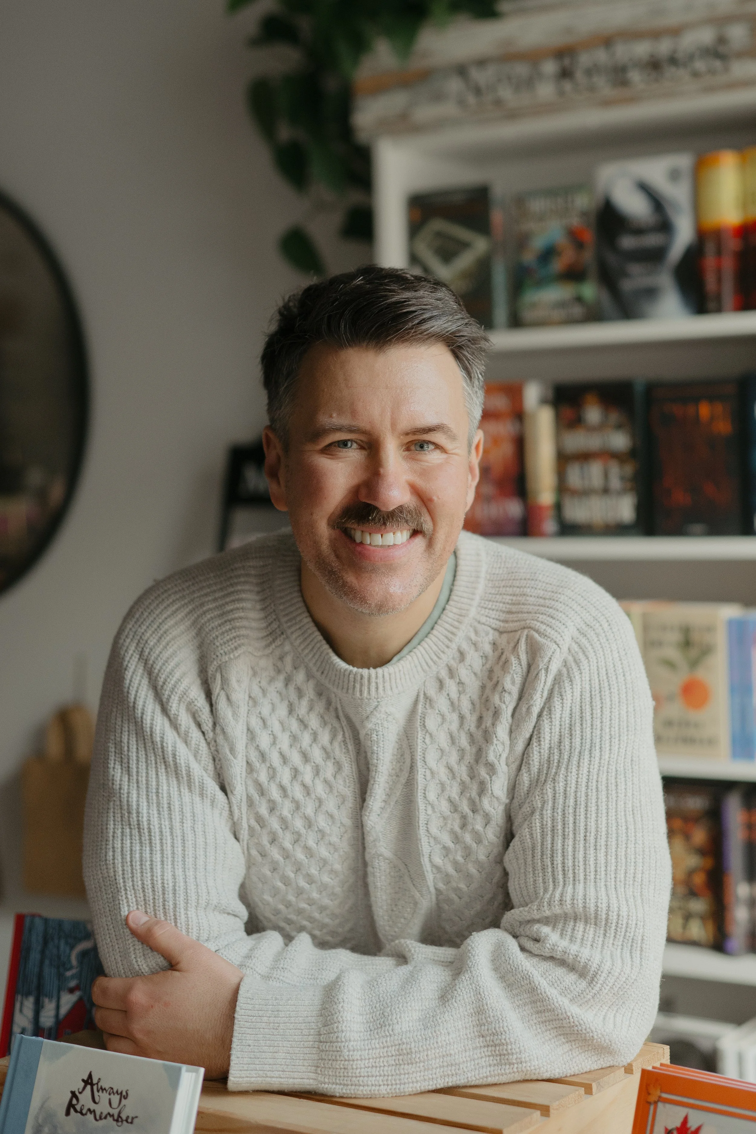 author Aaron Kyte in cream coloured sweater, smiling at the camera. Out of focus books in the background.