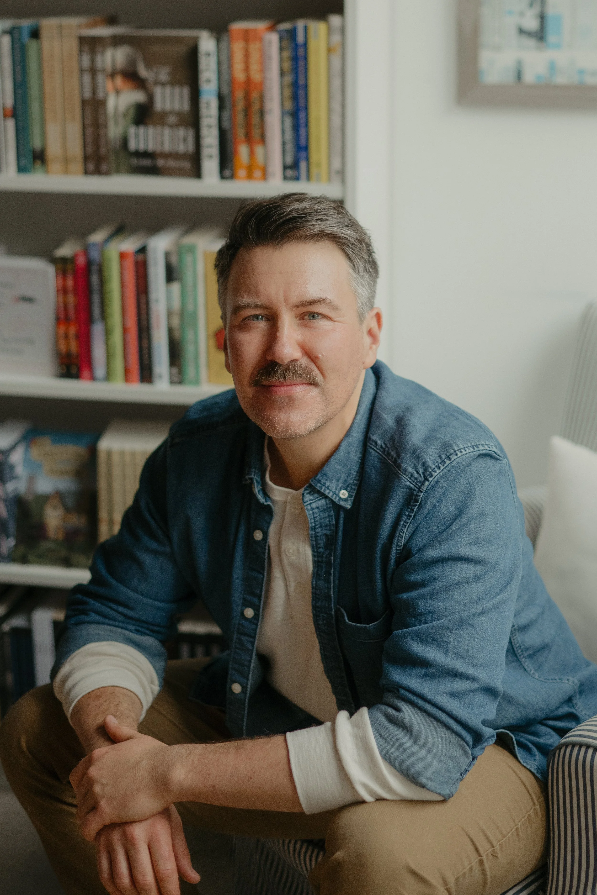Author Aaron Kyte sits, leaned forward, in a chair. He is wearing a blue denim shirt, unbuttoned over a cream-coloured henley, with tan slacks. Behind him are shelves of books out of of focus.