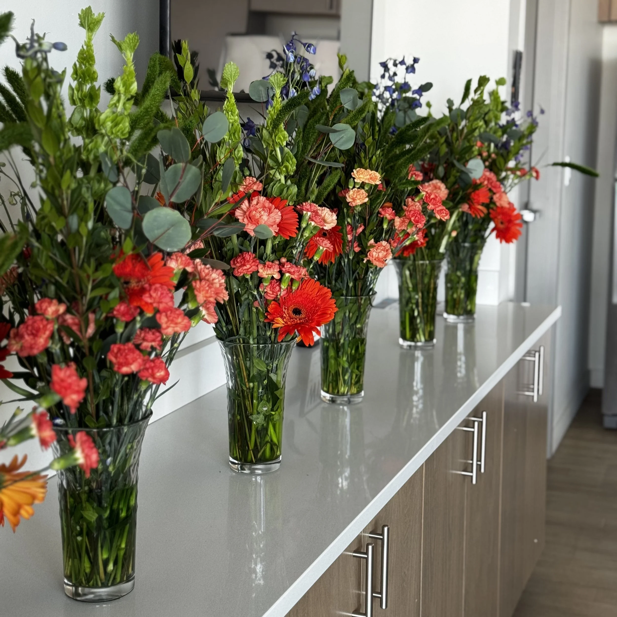 Five vase filled with orange gerbera daisies, orange carnations, blue eucalyptus and green millet on a clubhouse countertop at an event.