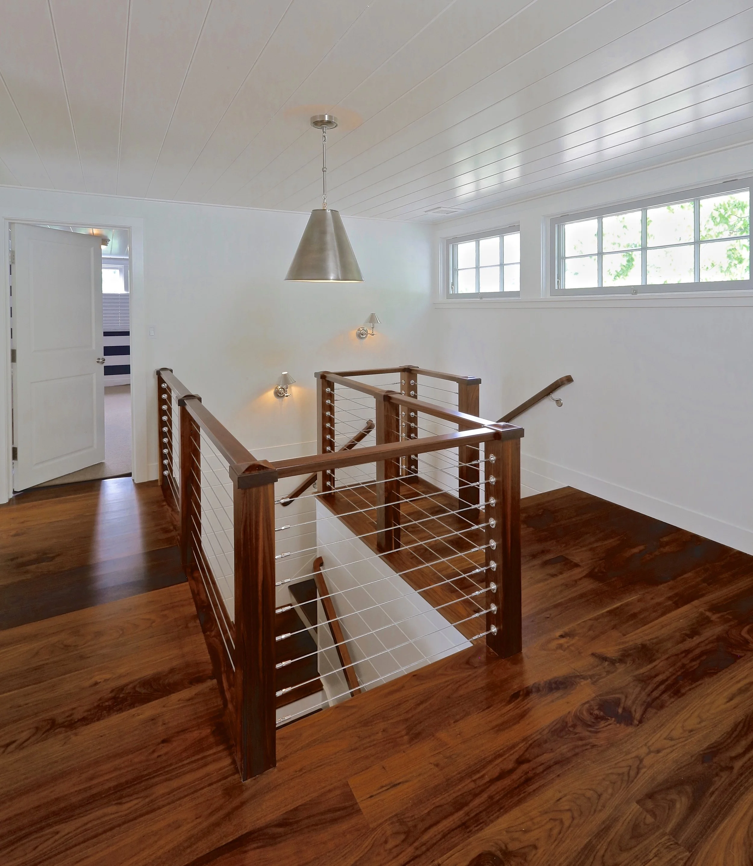 Interior view of a staircase landing with wooden flooring, a metal and wood staircase railing, white walls, three horizontal windows, and a ceiling with wood paneling, a modern pendant light, and wall-mounted lights.