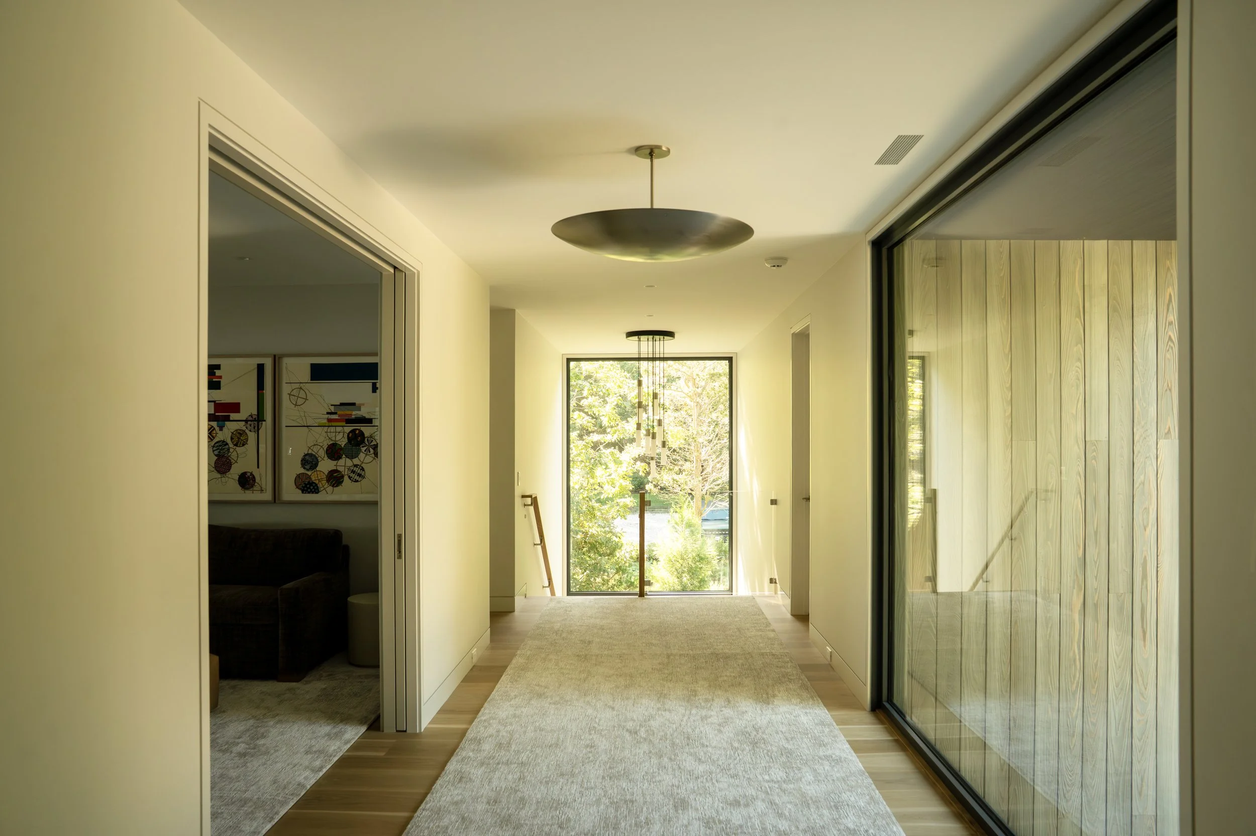 Bright corridor with large window at the end, modern lighting fixtures, glass sliding doors on the right, minimalist decor, and wooden flooring.