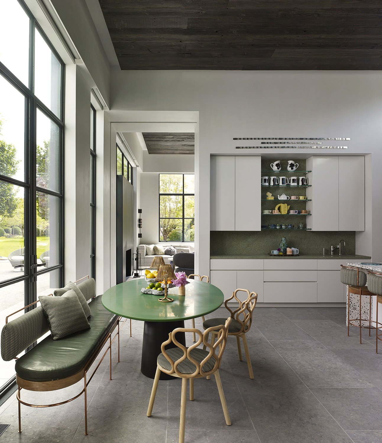 Modern kitchen and dining area with a green oval table, beige sofa, and large windows showing an outdoor view.