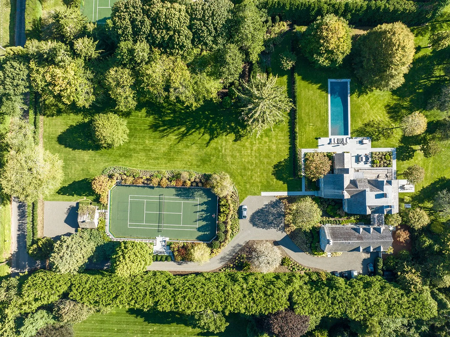 Aerial view of a large backyard with a tennis court, a swimming pool, and a house surrounded by trees and greenery.