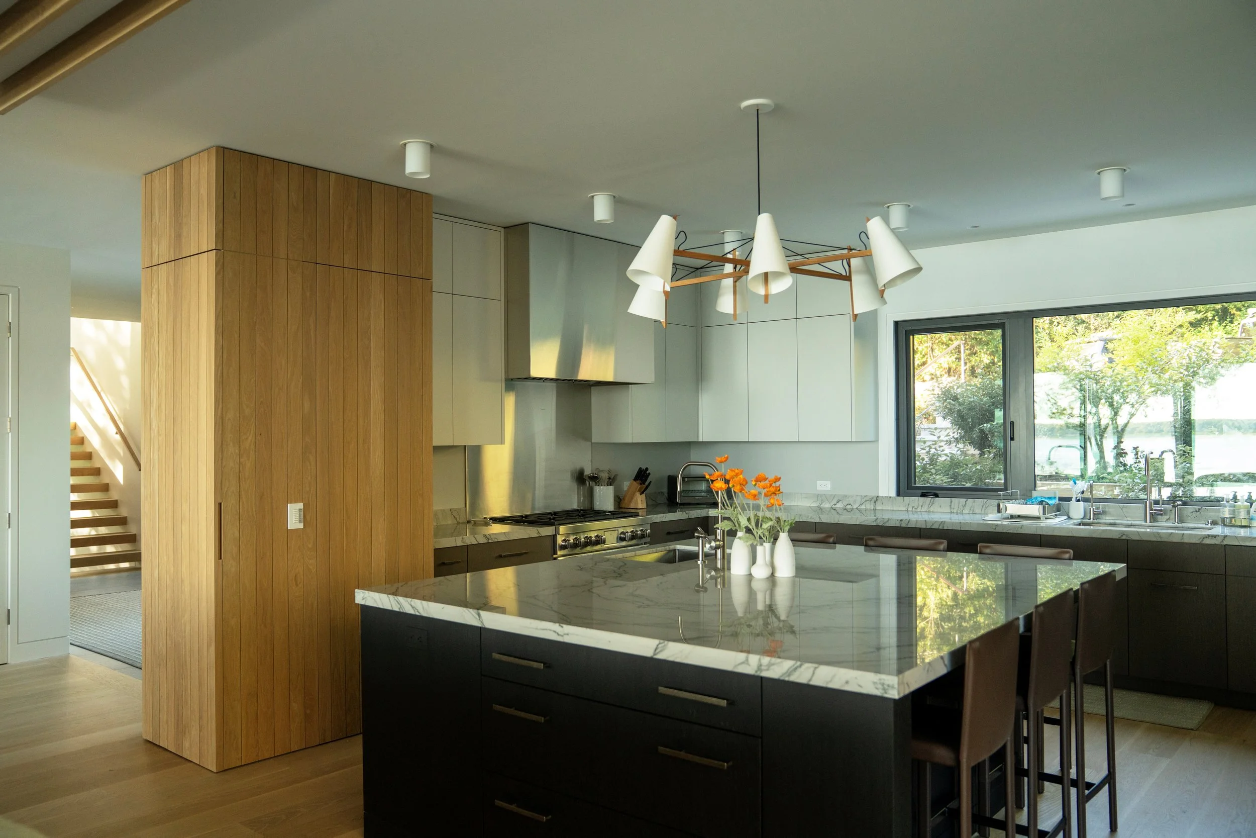 Modern kitchen with black island, marble countertops, wooden accents, and large window overlooking greenery.