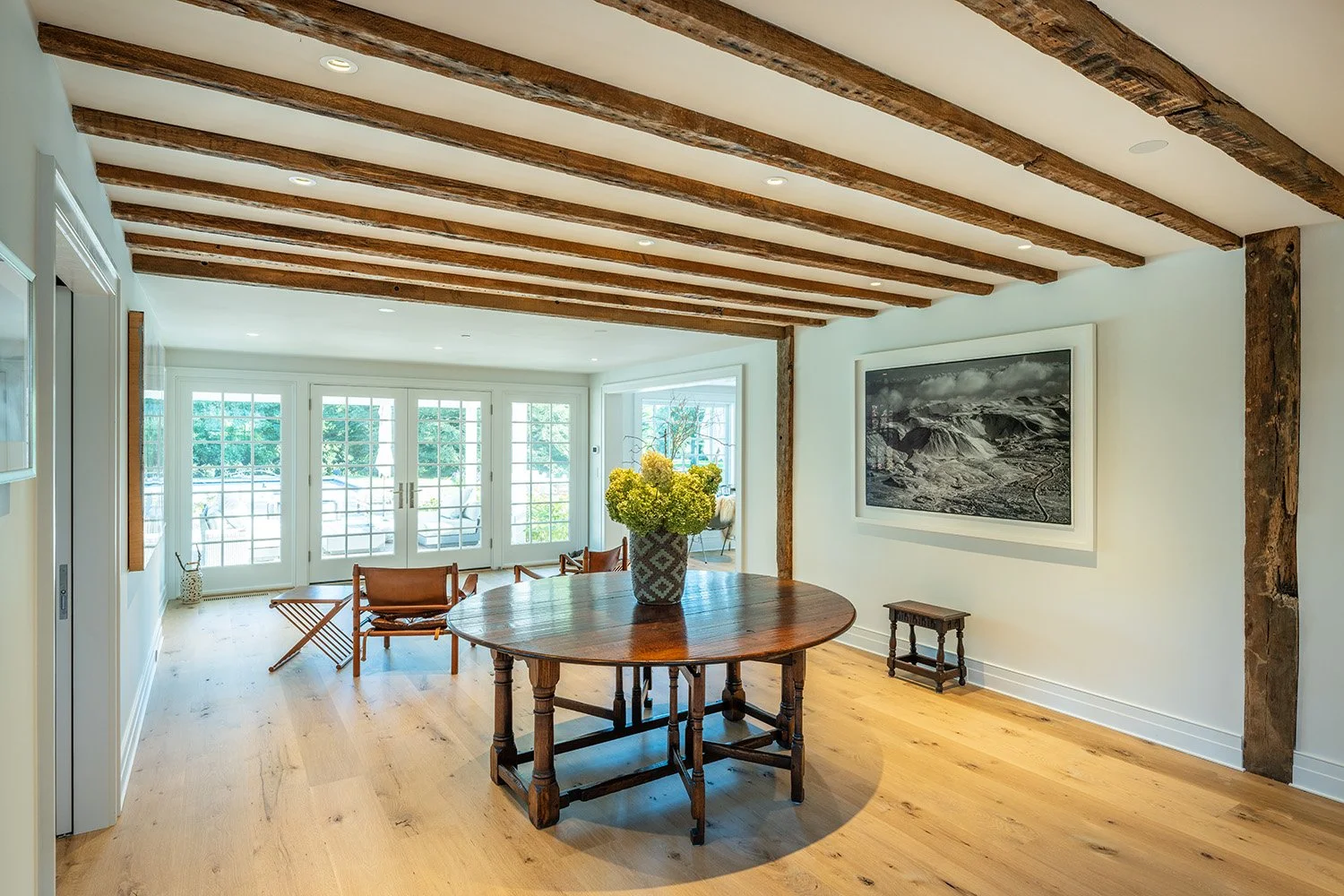 Bright dining room with wooden beams on the ceiling, a round wooden table with a vase of yellow flowers, and large windows with doors leading to an outdoor space.