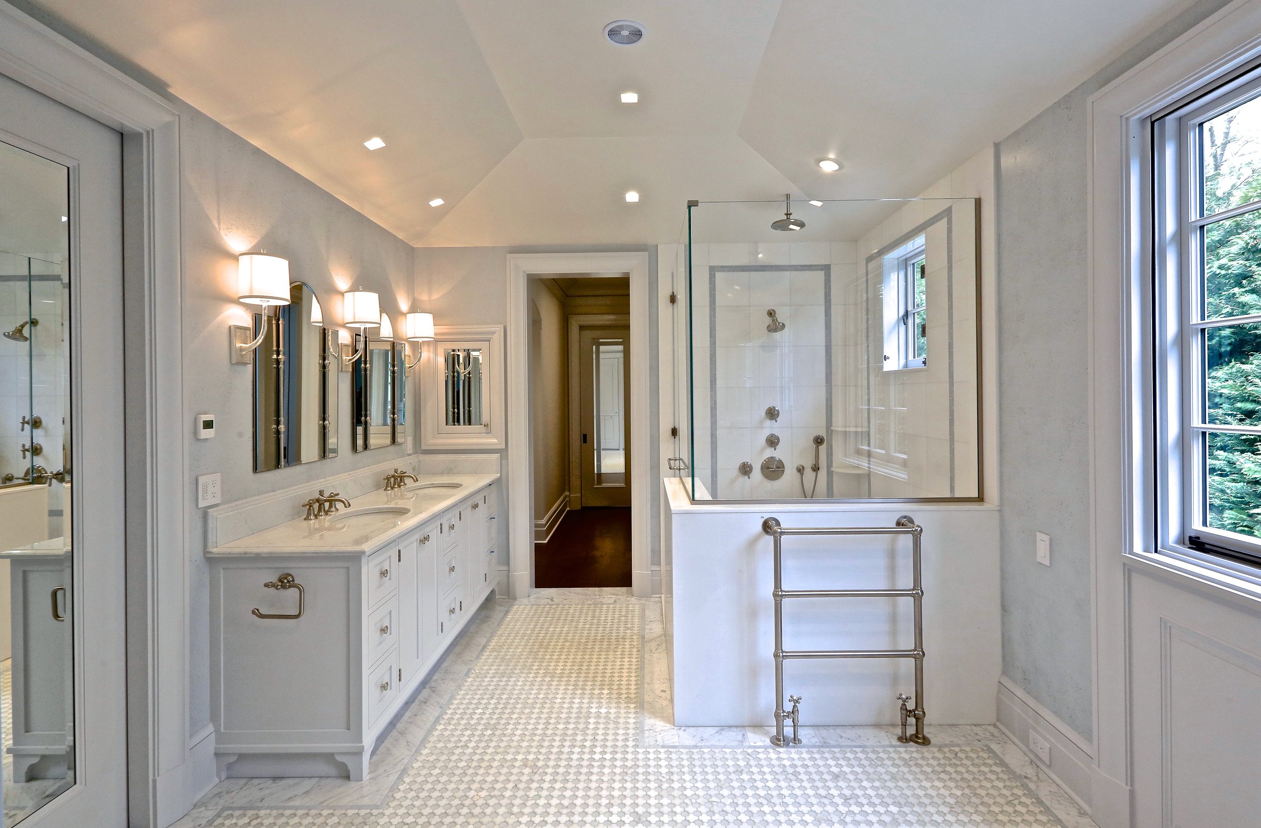 Bright and elegant bathroom with a double vanity, three mirrors, wall-mounted lights, a glass-enclosed shower with multiple shower heads, a window with greenery outside, and a heated towel rack.