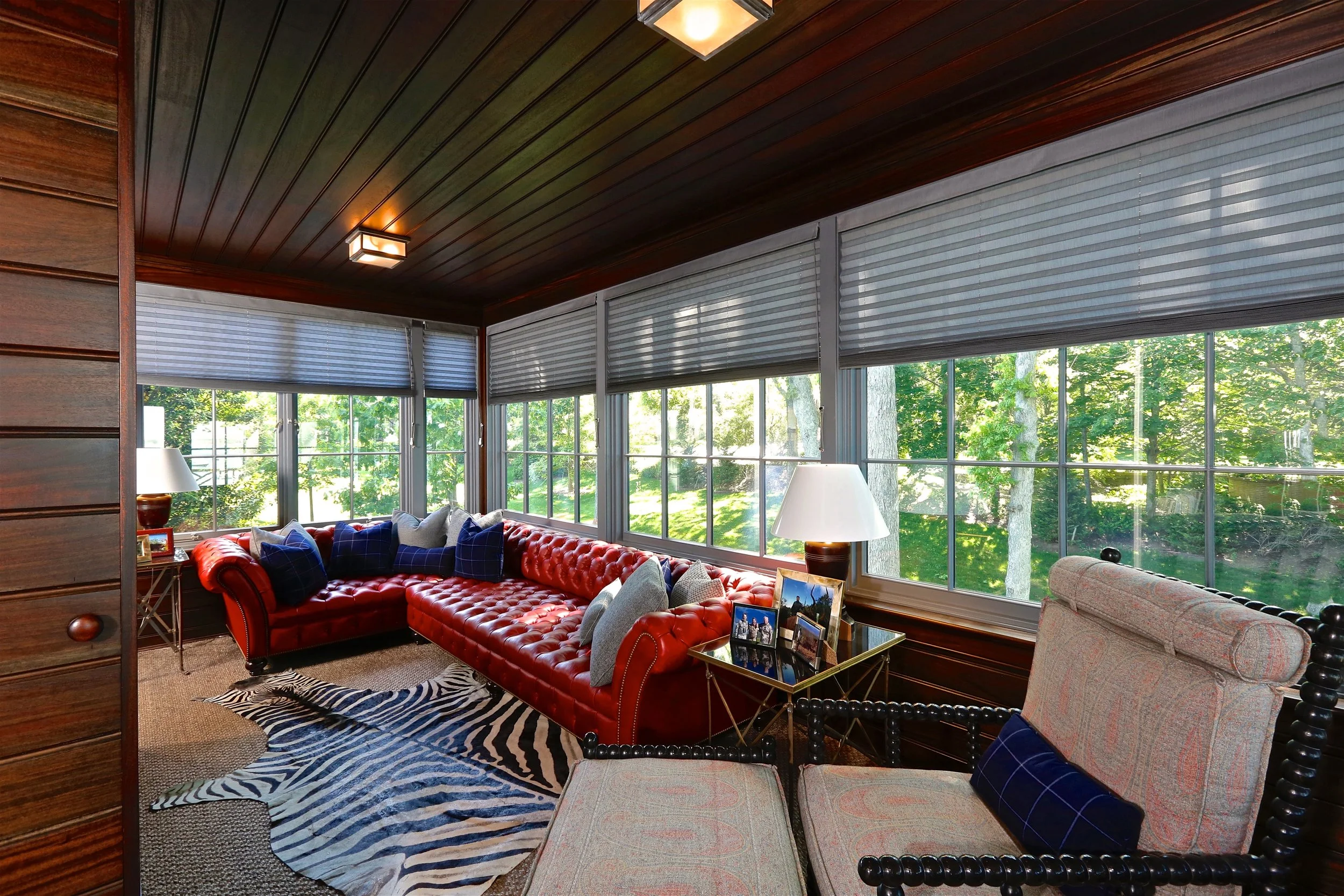 Sunroom with large windows, red leather sectional sofa, patterned area rug, side tables with lamps, and view of green trees outside.