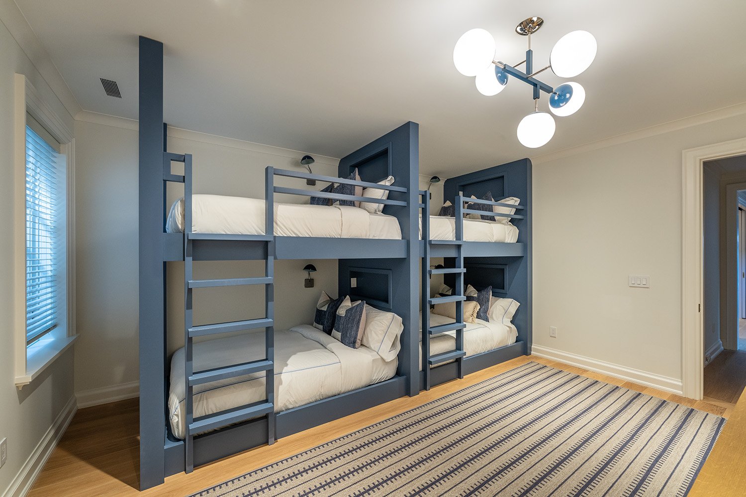 Room with two bunk beds painted blue, each with white bedding and dark decorative pillows. A large window with blinds is on the left, and a striped rug covers the wooden floor. Modern ceiling light fixture and wall-mounted reading lights are present.