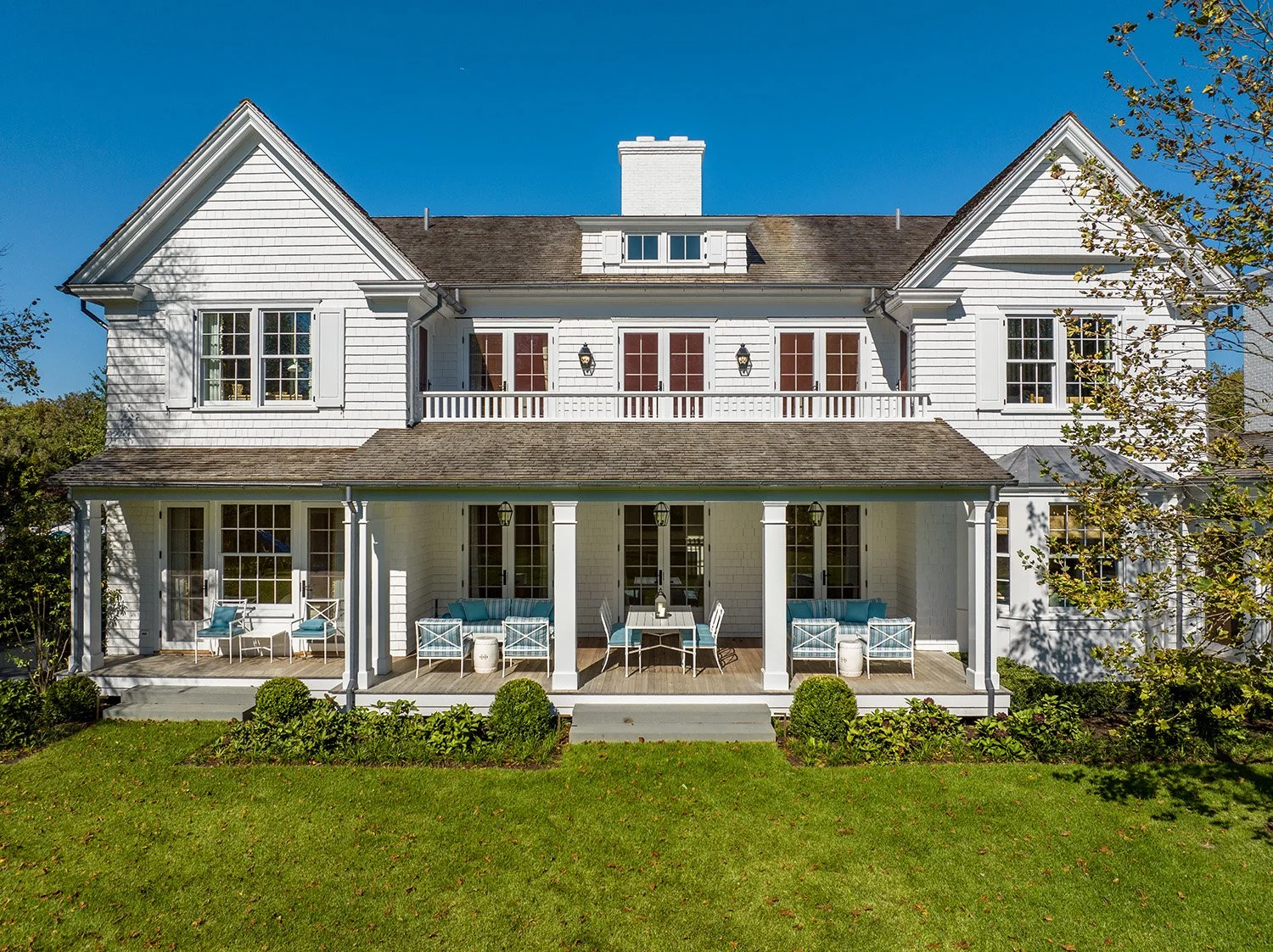A large white two-story house with a covered porch, multiple windows with shutters, and a balcony on the second floor. The house has a pitched roof with a chimney and is surrounded by a well-kept lawn with bushes and trees.