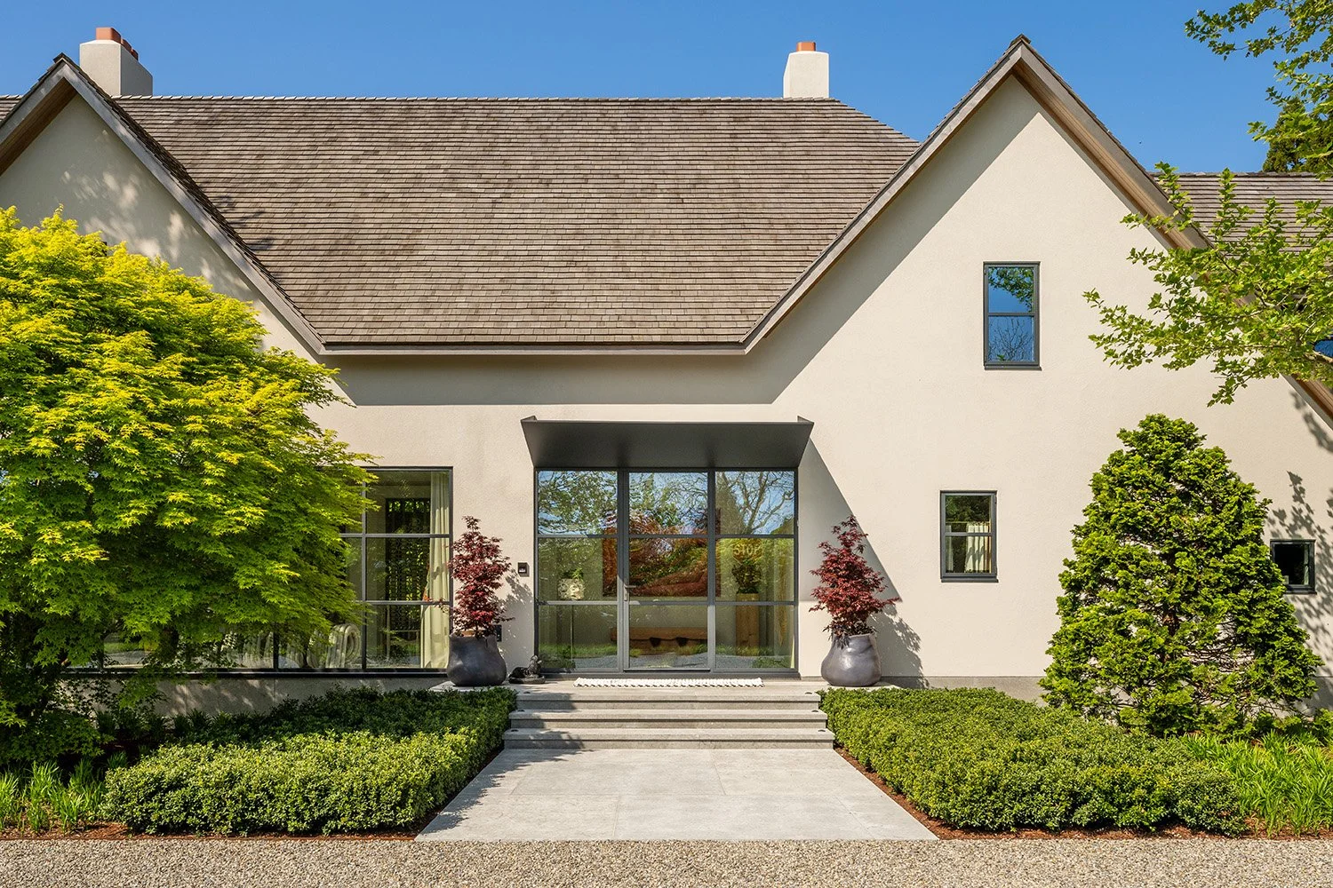 Front view of a modern house with a gabled roof, white exterior walls, multiple windows, and a glass front door, surrounded by lush green trees and shrubs under a clear blue sky.