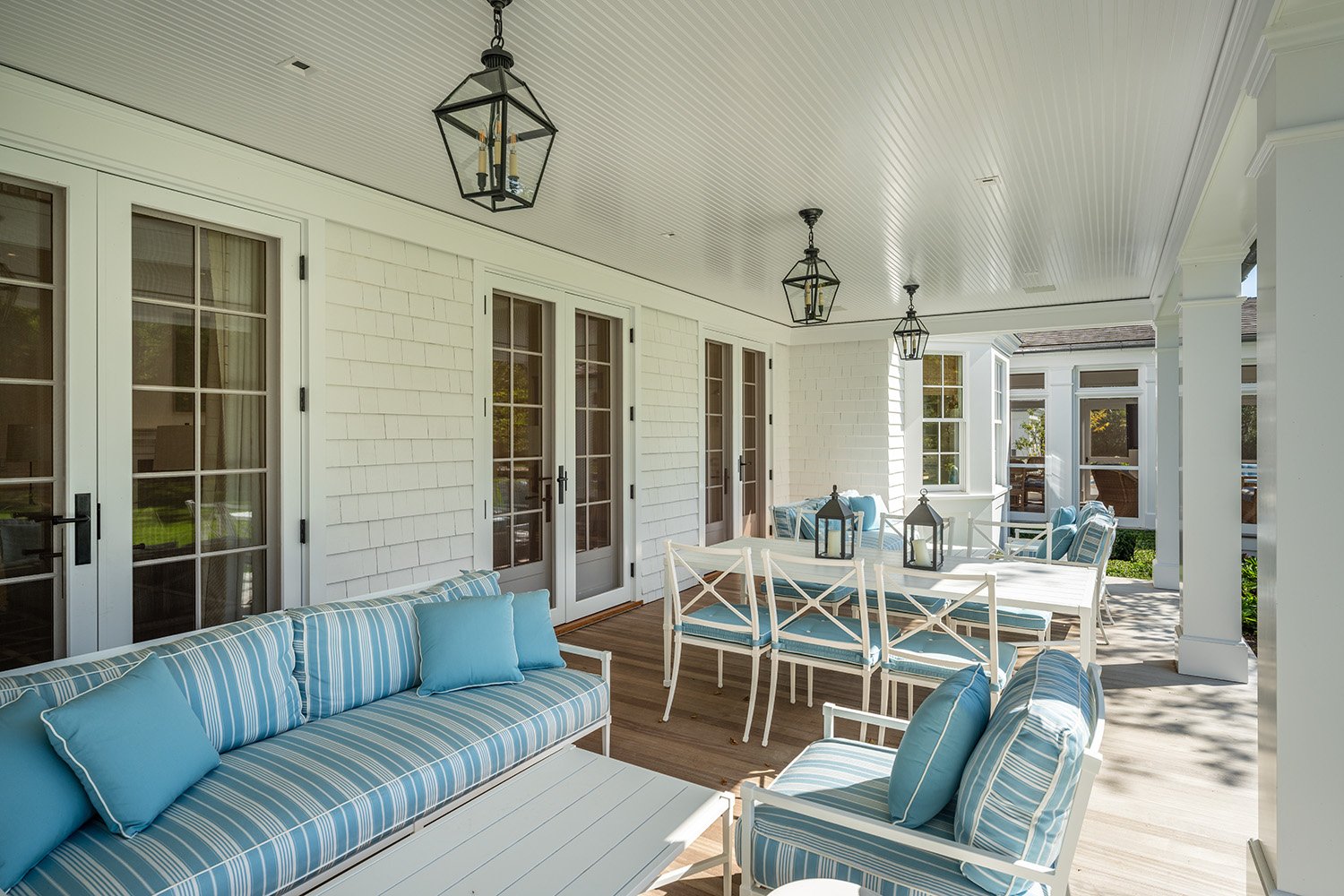 Covered porch with white furniture, blue-striped cushions, black lantern-style hanging lights, and white exterior walls with glass door windows.
