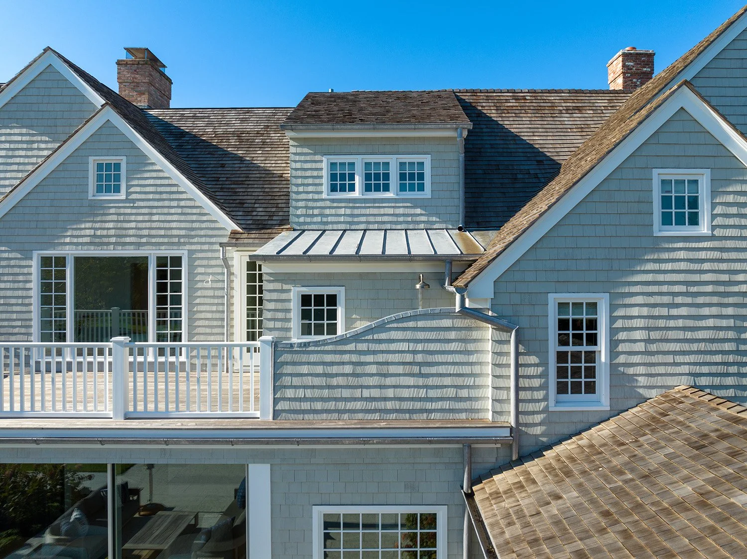 Rear view of a house with a balcony, multiple windows, shingle siding, and a metal roof section, under a clear blue sky.