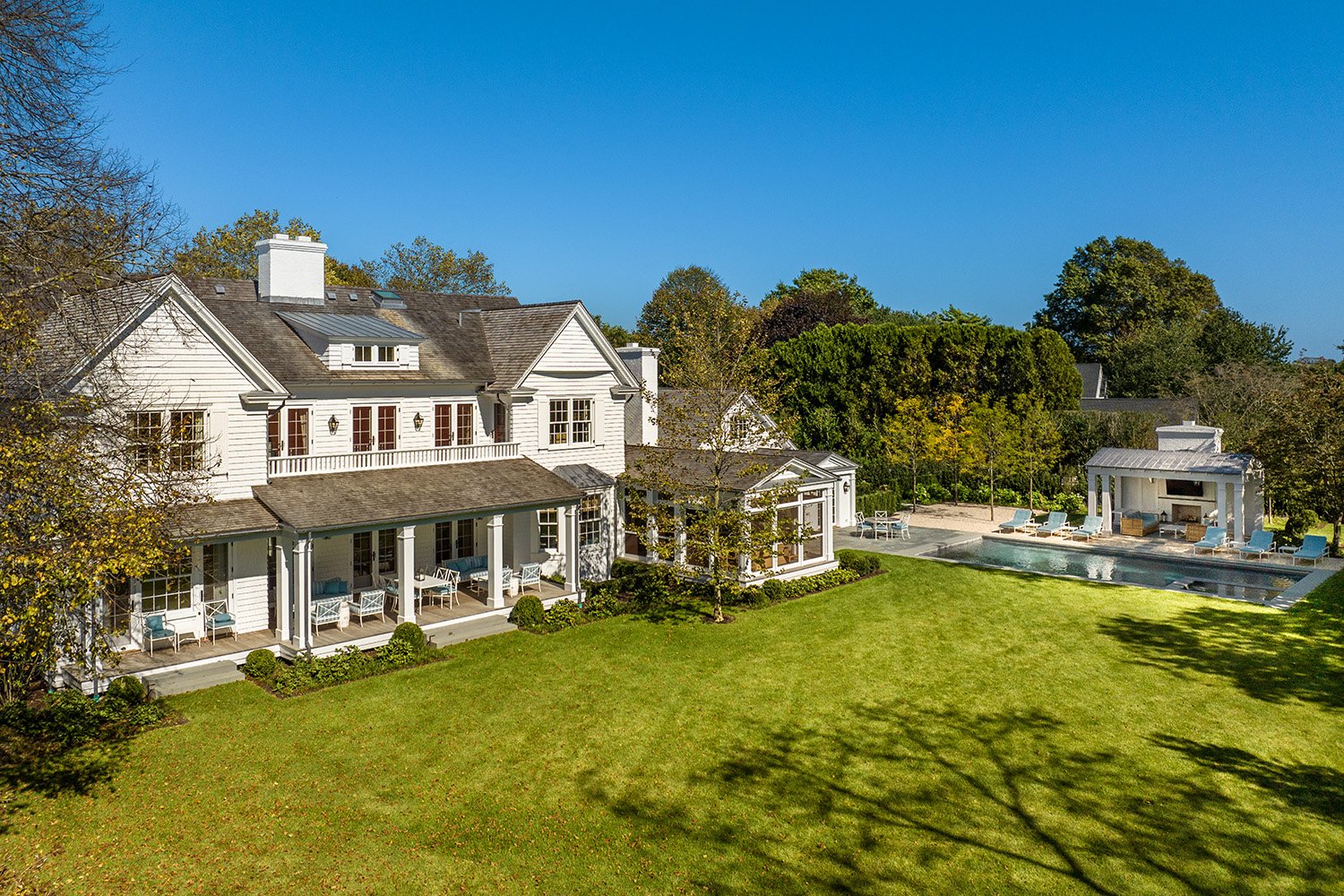 A large white house with multiple stories, a porch with white furniture, and a backyard with a lawn, trees, and a swimming pool with a shaded seating area.
