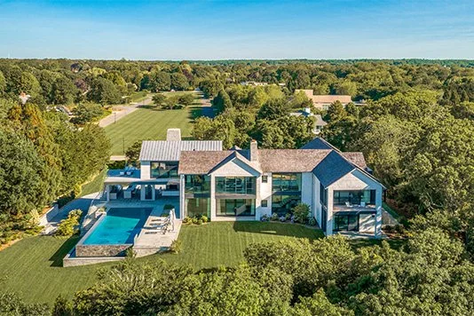 A modern house with an outdoor pool, surrounded by lush green lawn and trees, located in a spacious rural area with a sports field in the background.