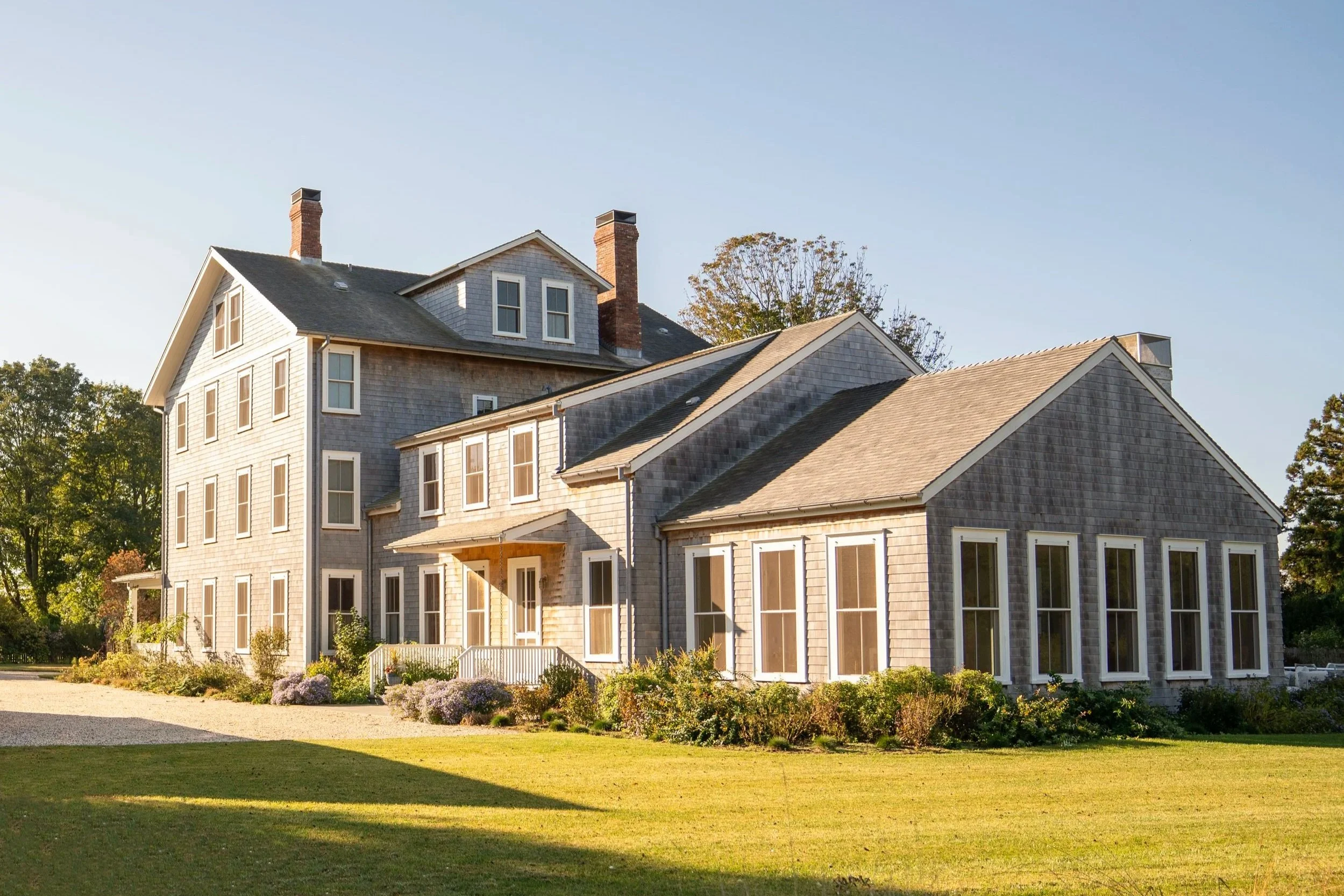 Hamptons large multi-story house with gray shingle siding, multiple windows, and a landscaped front yard, under a clear blue sky.
