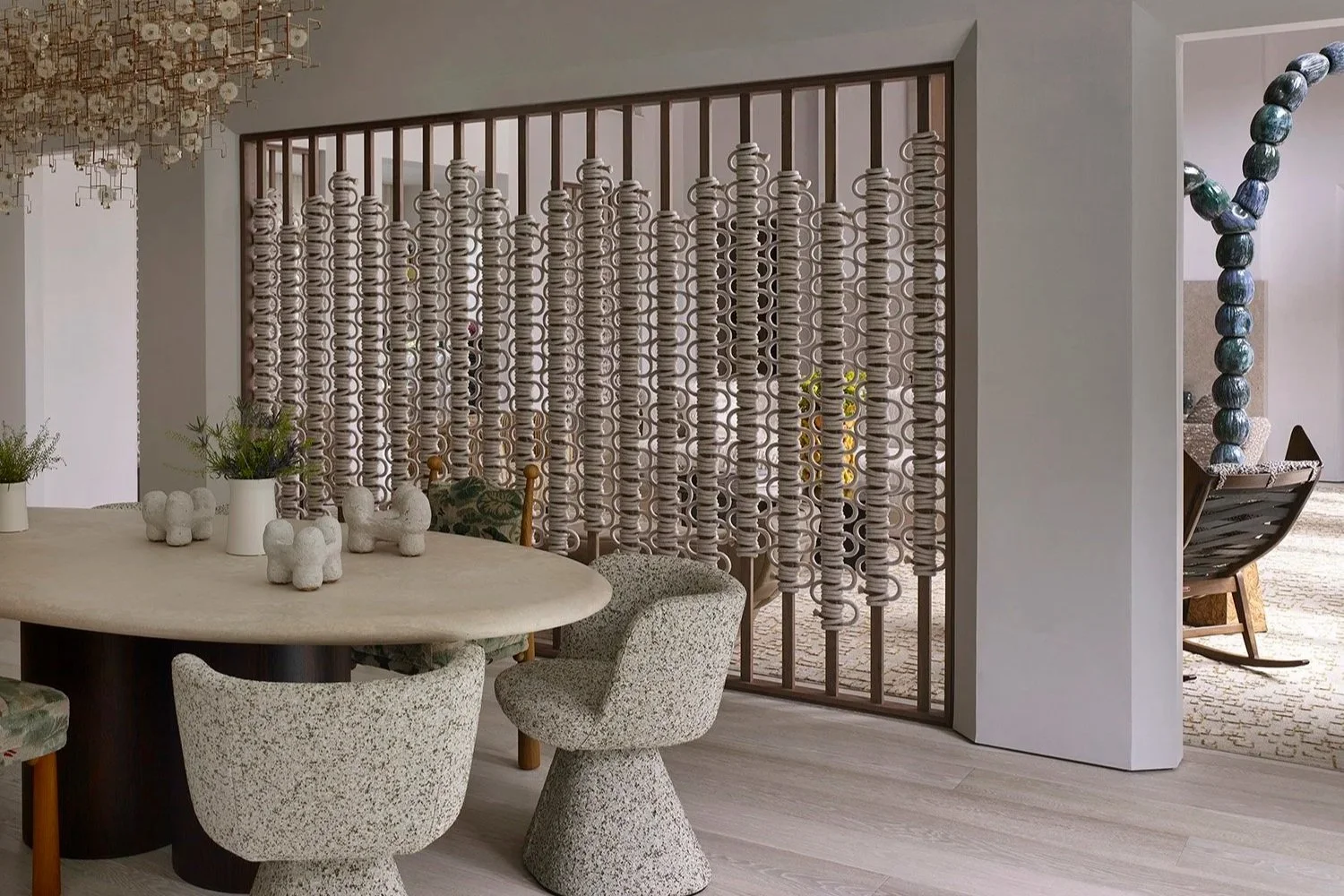 Contemporary dining area with beige speckled chairs around a beige table, decorated with white vases and plants, and a decorative partition with hanging loops made of wood and woven rings, with a glimpse of a cozy sitting nook with a rocking chair an