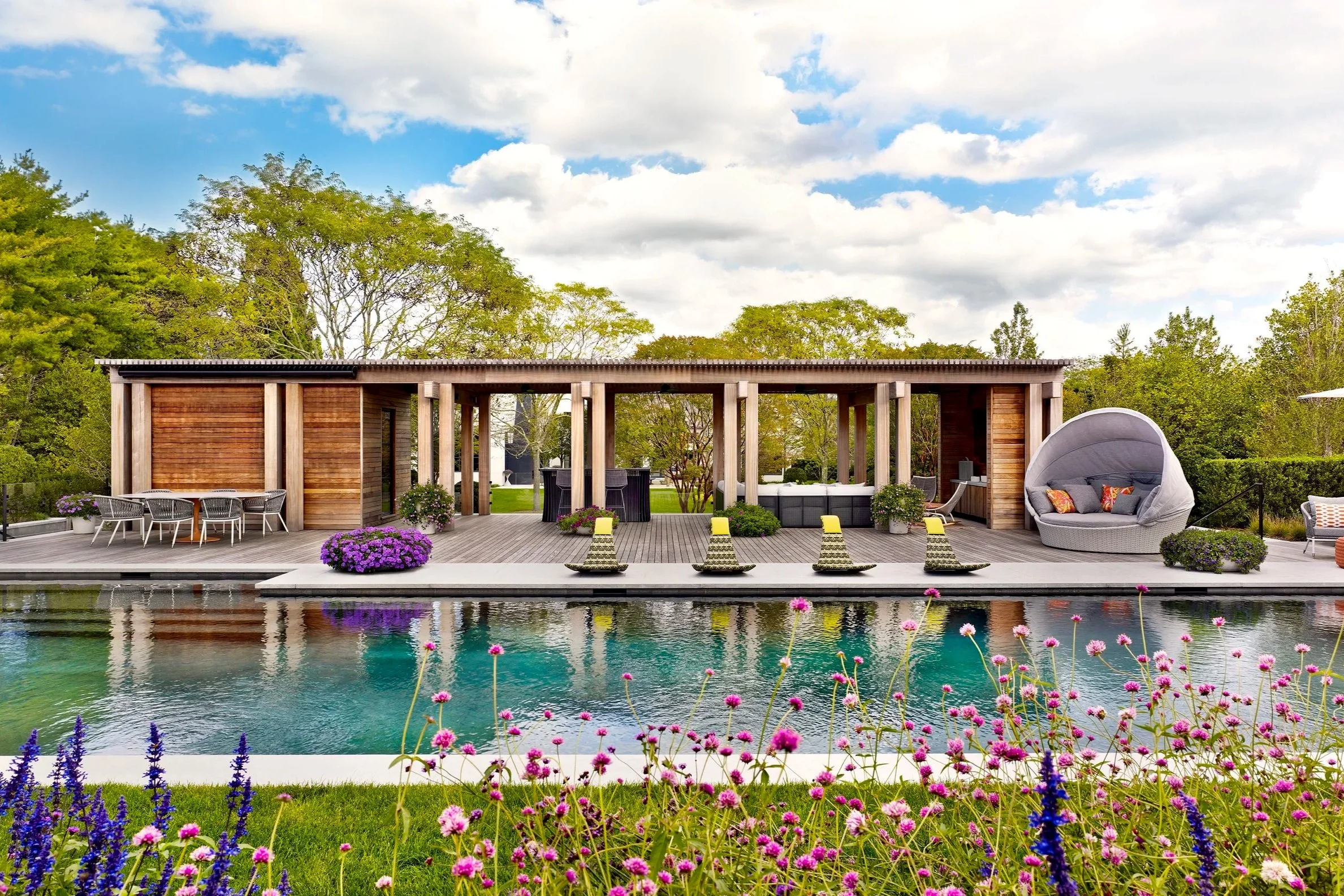 Modern backyard with a swimming pool, colorful flowers in the foreground, a wooden deck, and outdoor furniture including a round lounge chair with cushions, a dining table with chairs, and a covered sitting area with plants and umbrellas, surrounded 