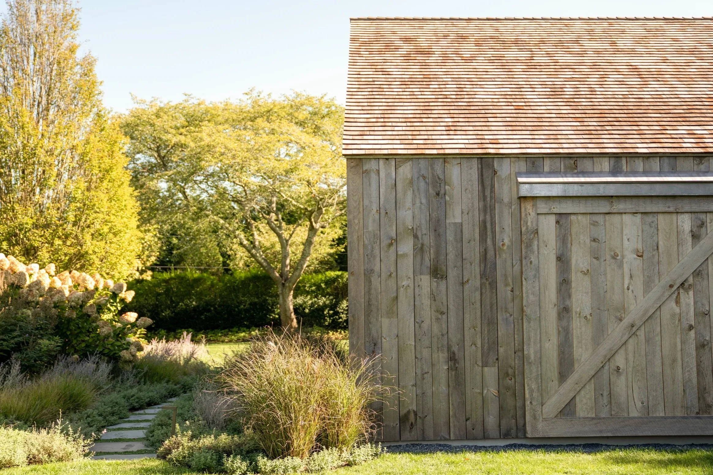 A wooden shed with a sloped roof in a garden, surrounded by various plants and trees with green foliage. Construction in the Hamptons
