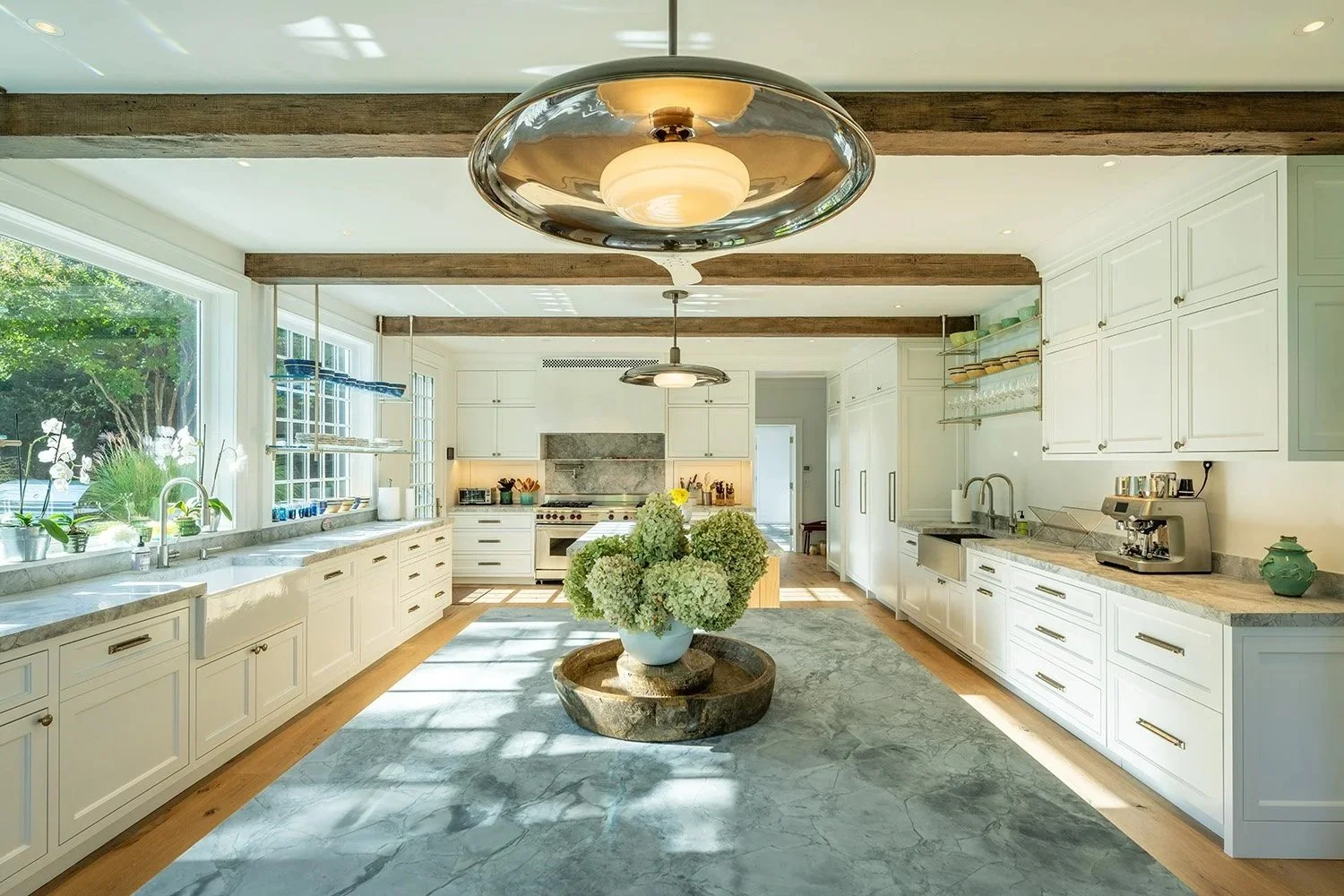 Bright kitchen with white cabinets, marble countertops, large window, wooden beams on ceiling, a round flower vase with hydrangeas in the center, and modern appliances. Bridgehampton New York.