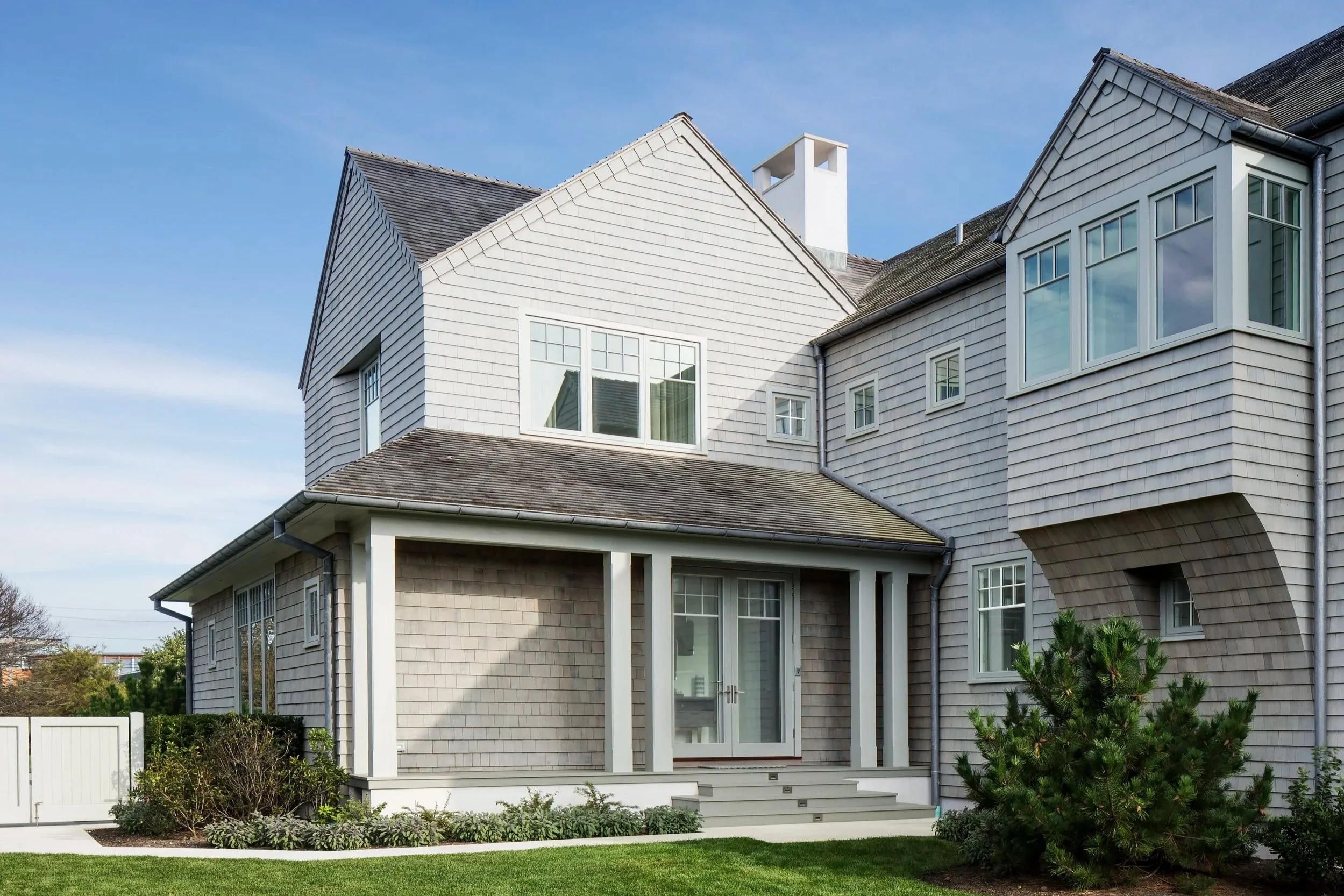 Exterior of a modern house with wooden shingles, a porch with steps, and a grassy yard, under a clear blue sky. Southampton custom design build. 