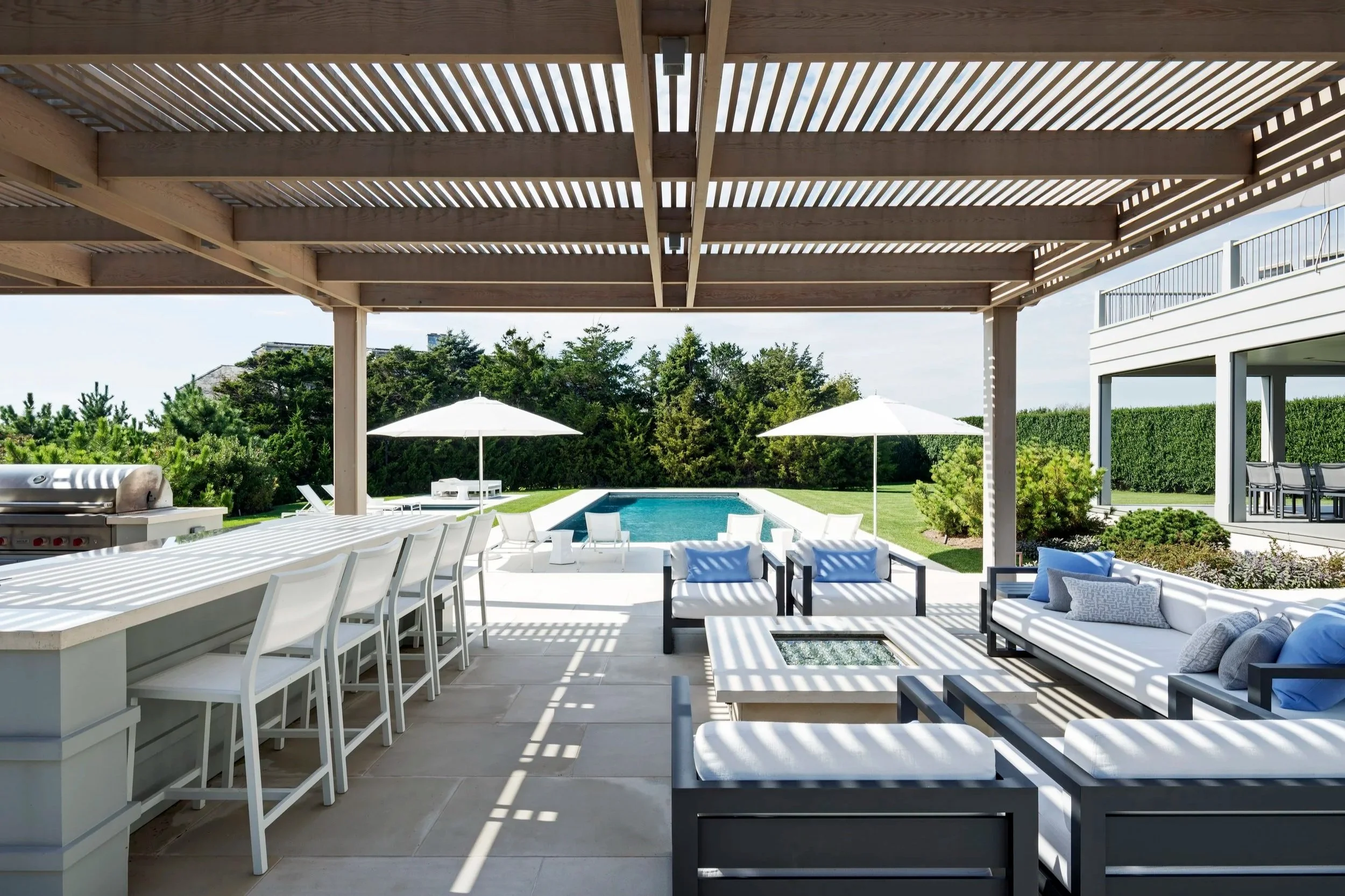 Patio area with white couches, glass-topped fire pit, bar seating at a counter, swimming pool with umbrellas, lounge chairs, surrounded by green lawns and bushes under a wooden pergola. East Hampton construction. 