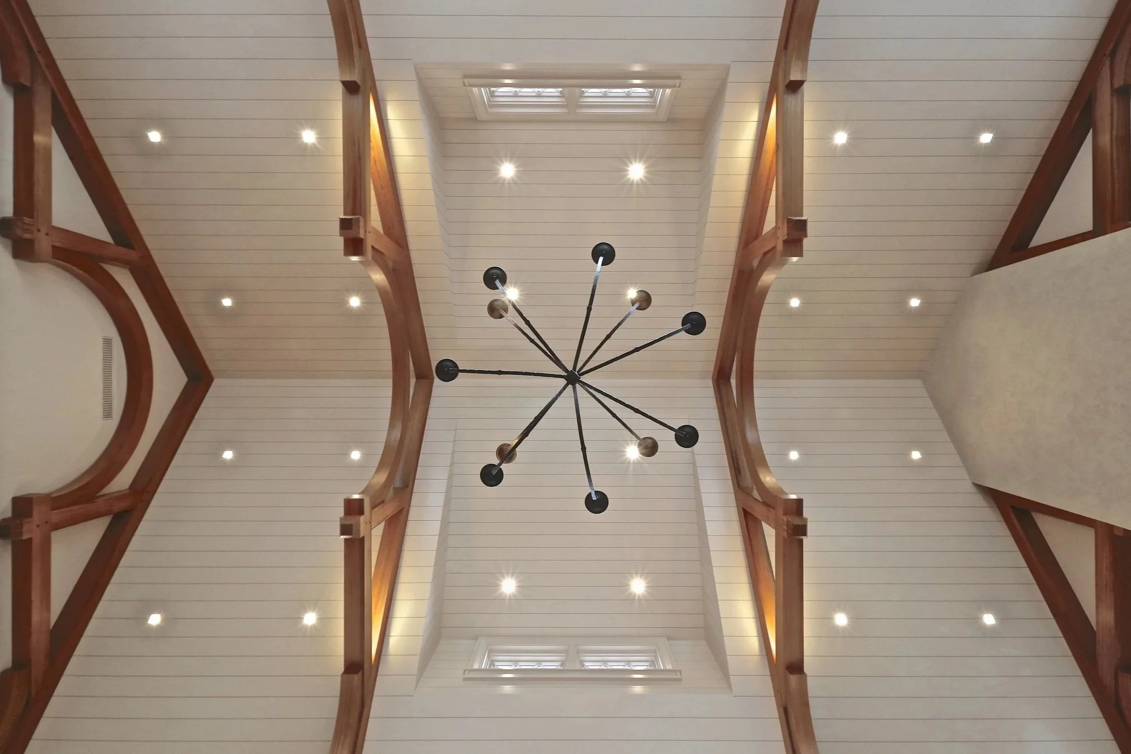 View of a high ceiling with a modern black chandelier, wooden beams, recessed lighting, and two windows at the top. Custom High End residential. 