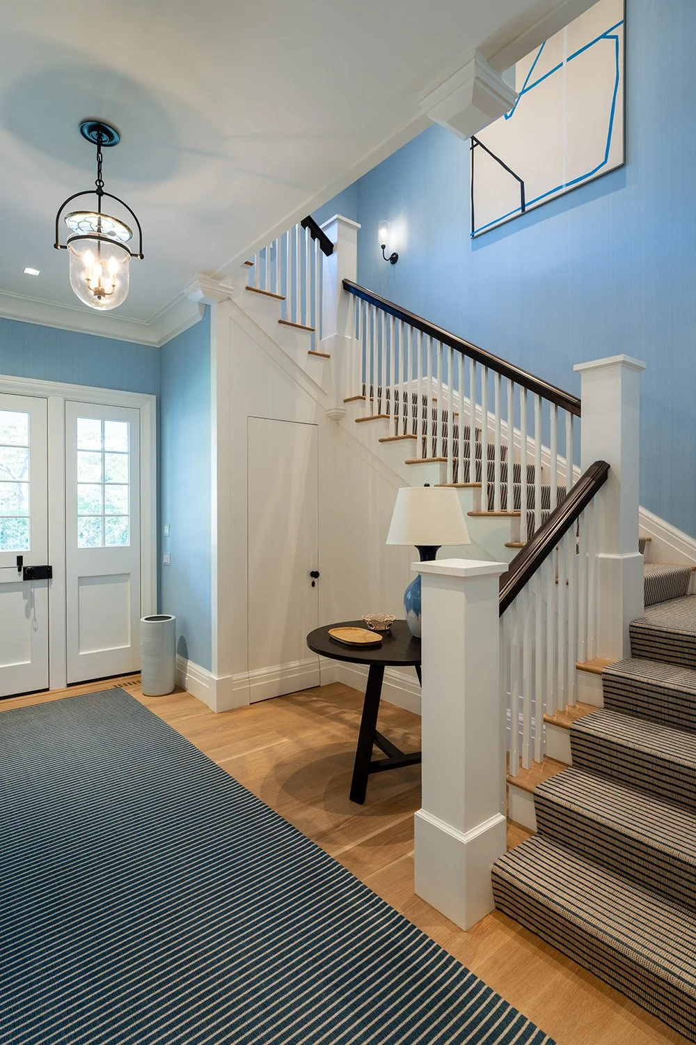 Entryway with blue walls, white staircase with dark handrail, striped carpet on stairs, black side table with lamp, painting, and decorative objects, front door with glass panes, light fixture hanging from ceiling.